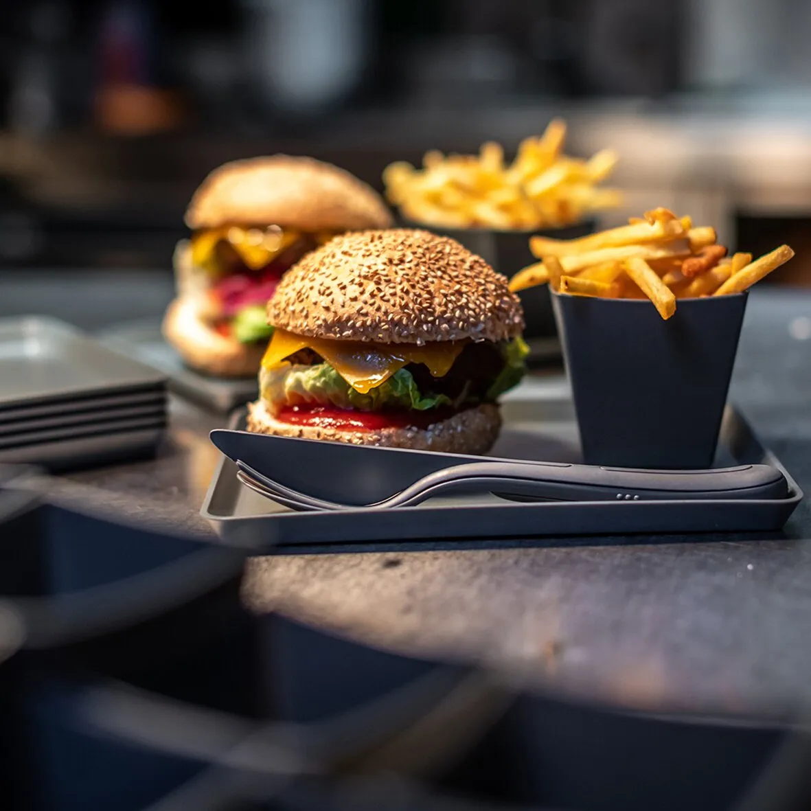 Graues Besteckset KLIKK mit Messer, Gabel und Löffel in einer Servierschale neben Burger und Pommes