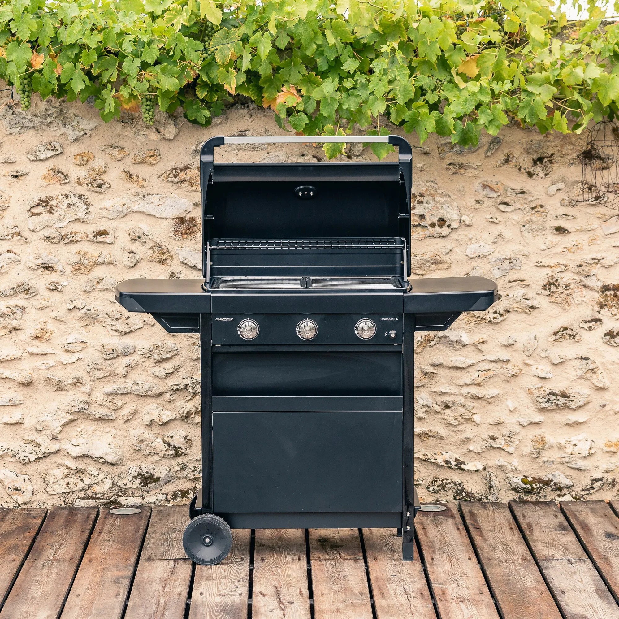 Schwarzer Gasgrill Compact 3 L mit geöffnetem Deckel, drei Bedienknöpfen und zwei Grillrosten auf Holzterrasse vor einer Weinrebe