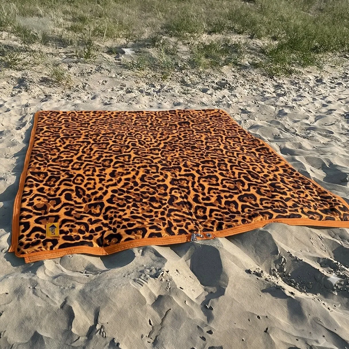 Auf Sand ausgebreiteter Vanlife-Schlafsack Bentcho im Leo-Design mit orangefarbenem Rand und Reißverschluss