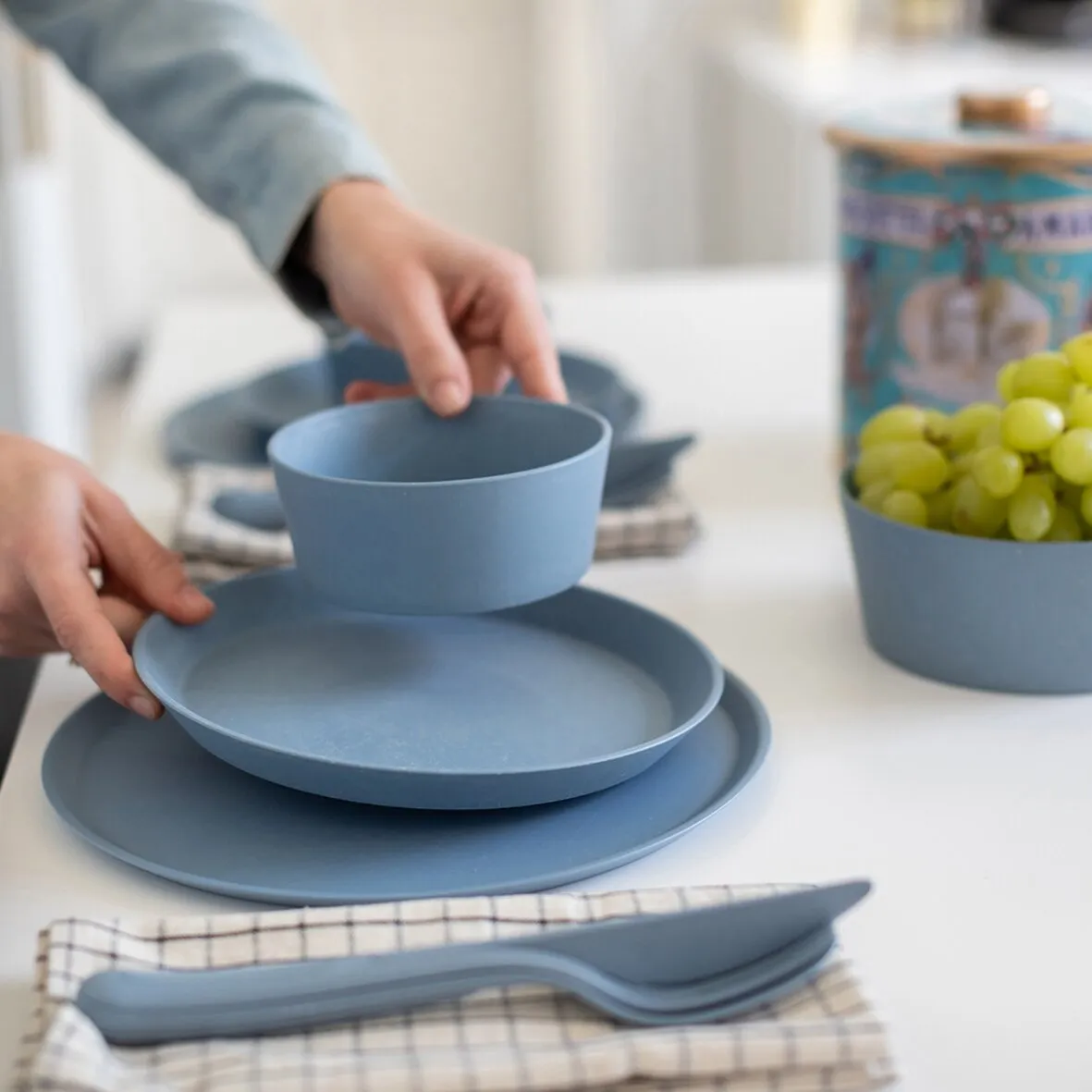Blaues KLIKK-Besteckset mit Gabel, Messer und Löffel auf kariertem Tuch neben blauem Geschirr und einer Schale Trauben