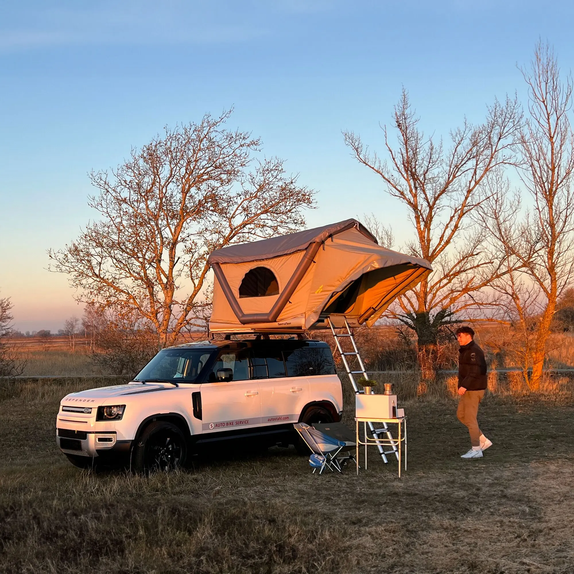 Dachzelt GT Roof in beige auf einem weißen SUV montiert, seitliche Leiter ausgeklappt, kahle Bäume und offenes Feld im Abendlicht