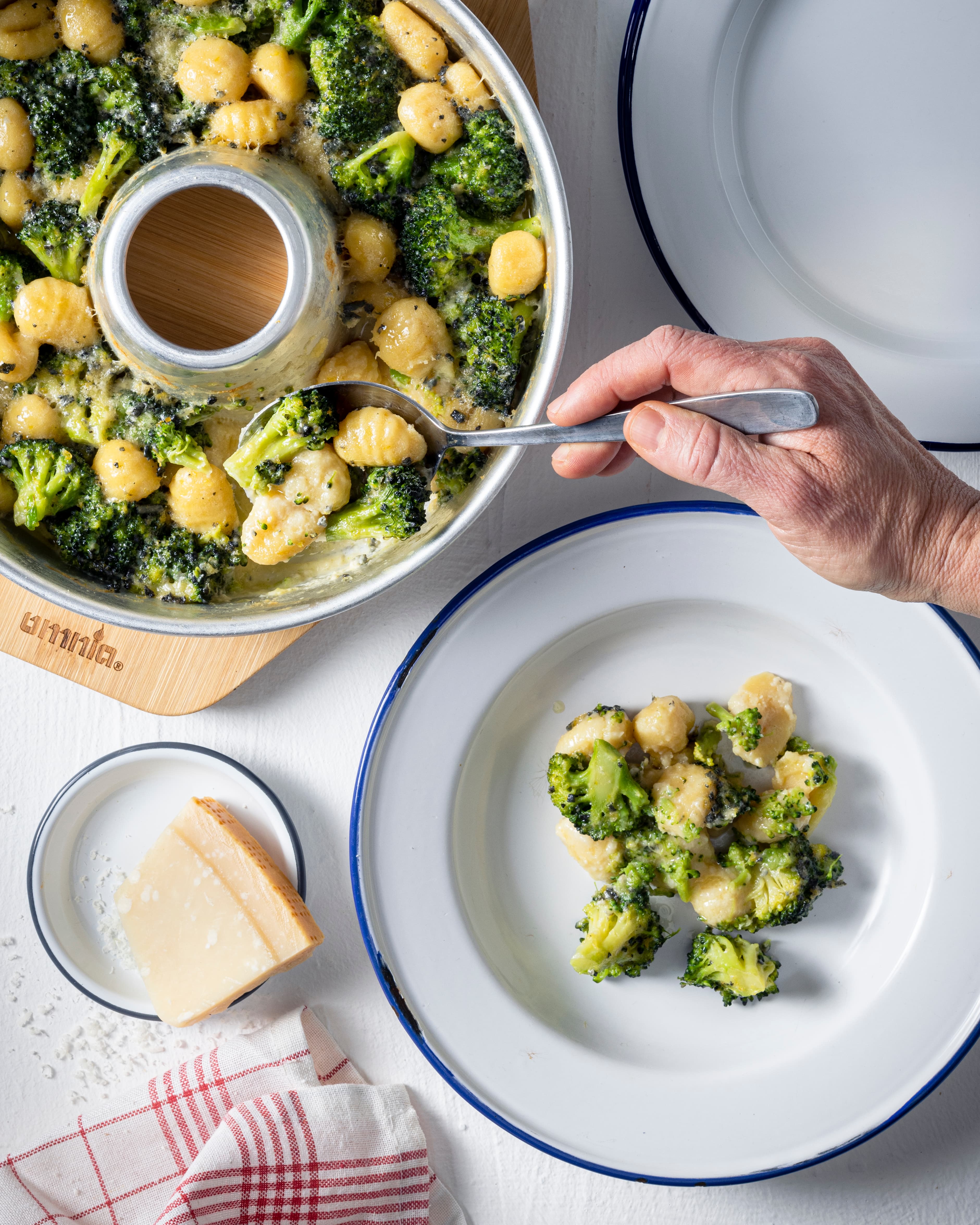 Draufsicht auf eine runde Kuchenform mit Rohrkern, gefüllt mit Brokkoli und Gnocchi in cremiger Sauce; eine Hand hält einen Löffel, Käsekeil und Teller daneben