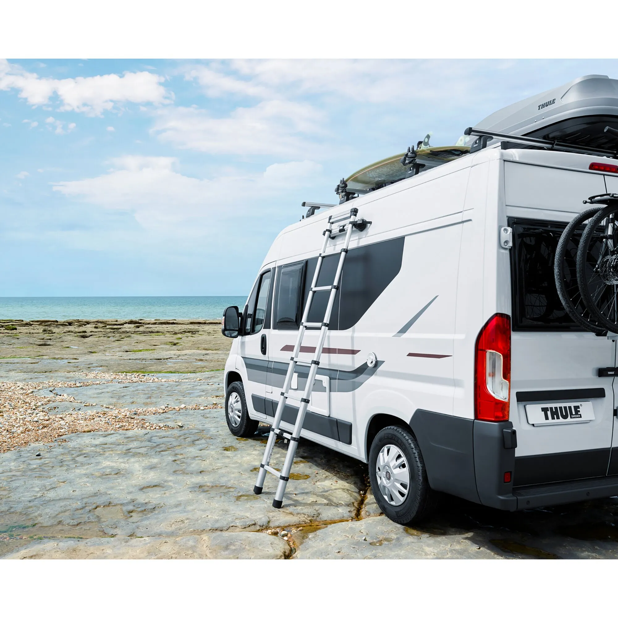 Thule Van Leiter aus Aluminium an der Seite eines weißen Wohnmobils am Meer, mit Dachbox, Surfbrettern und Fahrrädern ausgestattet