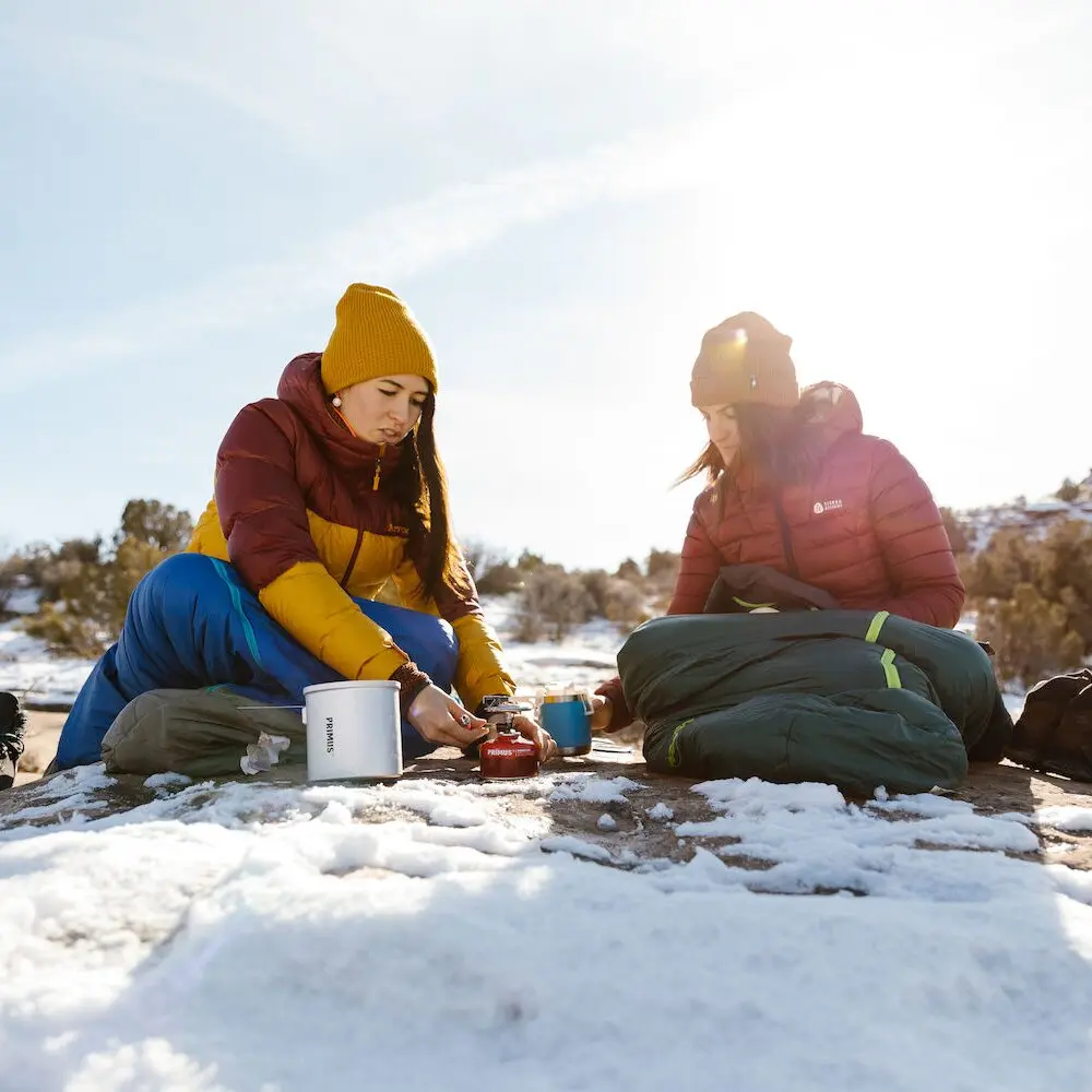 Zwei Personen sitzen im Schnee und kochen mit kleinem rotem Gaskocher, weißem Topf und blauem Becher vor sonnigem Winterhimmel
