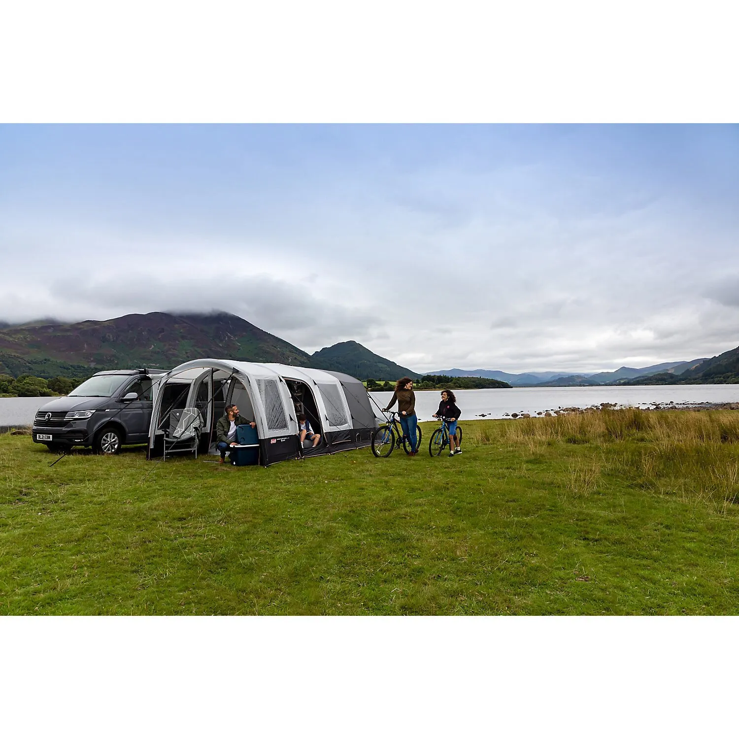 Graues Busvorzelt Journeymaster Deluxe L an einem Campervan mit geöffneten Fenstern, Menschen und Fahrrädern am Seeufer in Berglandschaft
