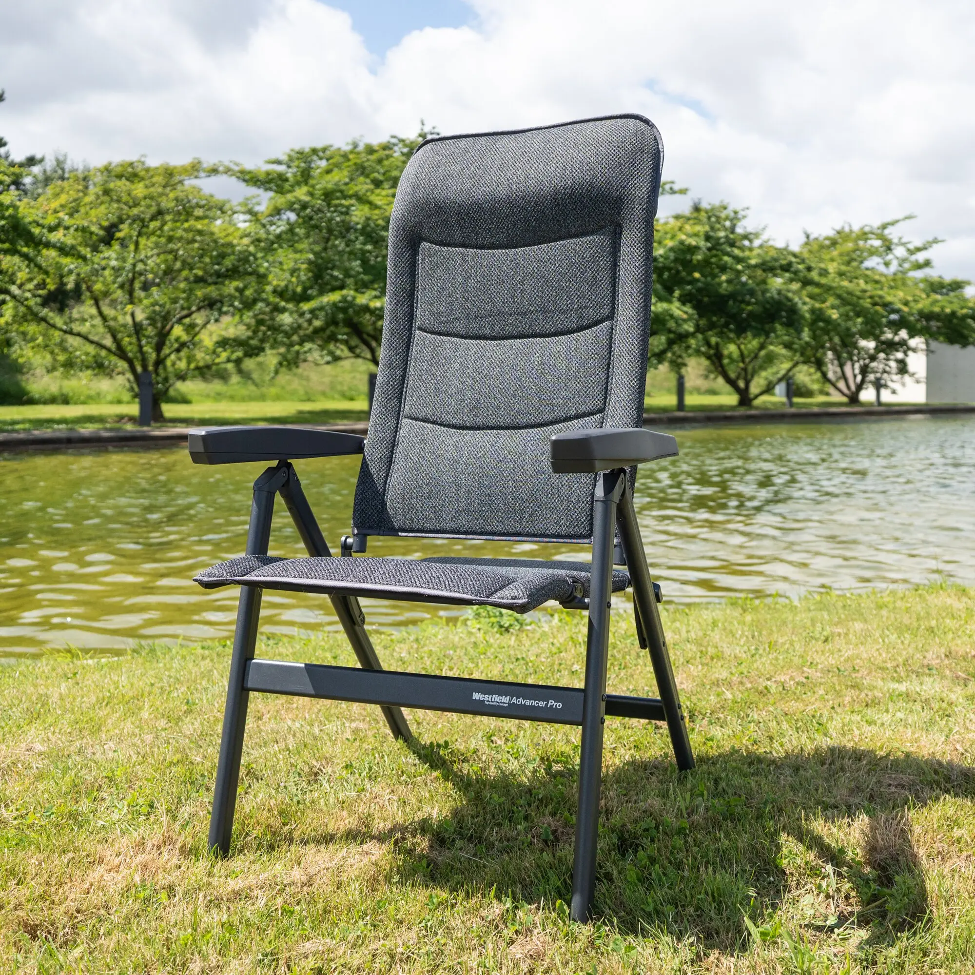 Rückenlehne eines dunkelgrauen, gewebten Campingstuhls Advancer Pro mit Armlehnen vor Gras, See und Bäumen in einer Outdoor-Szene