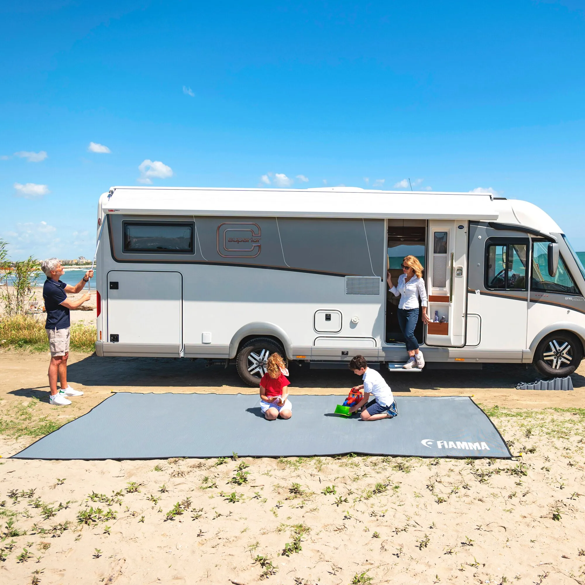 Grauer Fiamma Patio-Mat-Zeltteppich, 290 cm, vor einem Wohnmobil am Strand mit spielenden Kindern und Erwachsenen