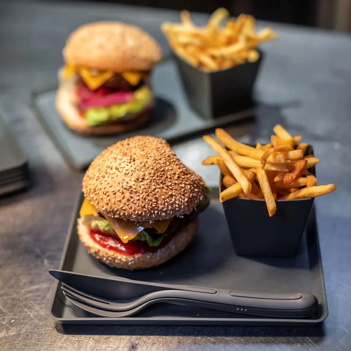 Graues Besteckset KLIKK mit Gabel und Messer auf einem Tablett neben Burgern und Pommes frites