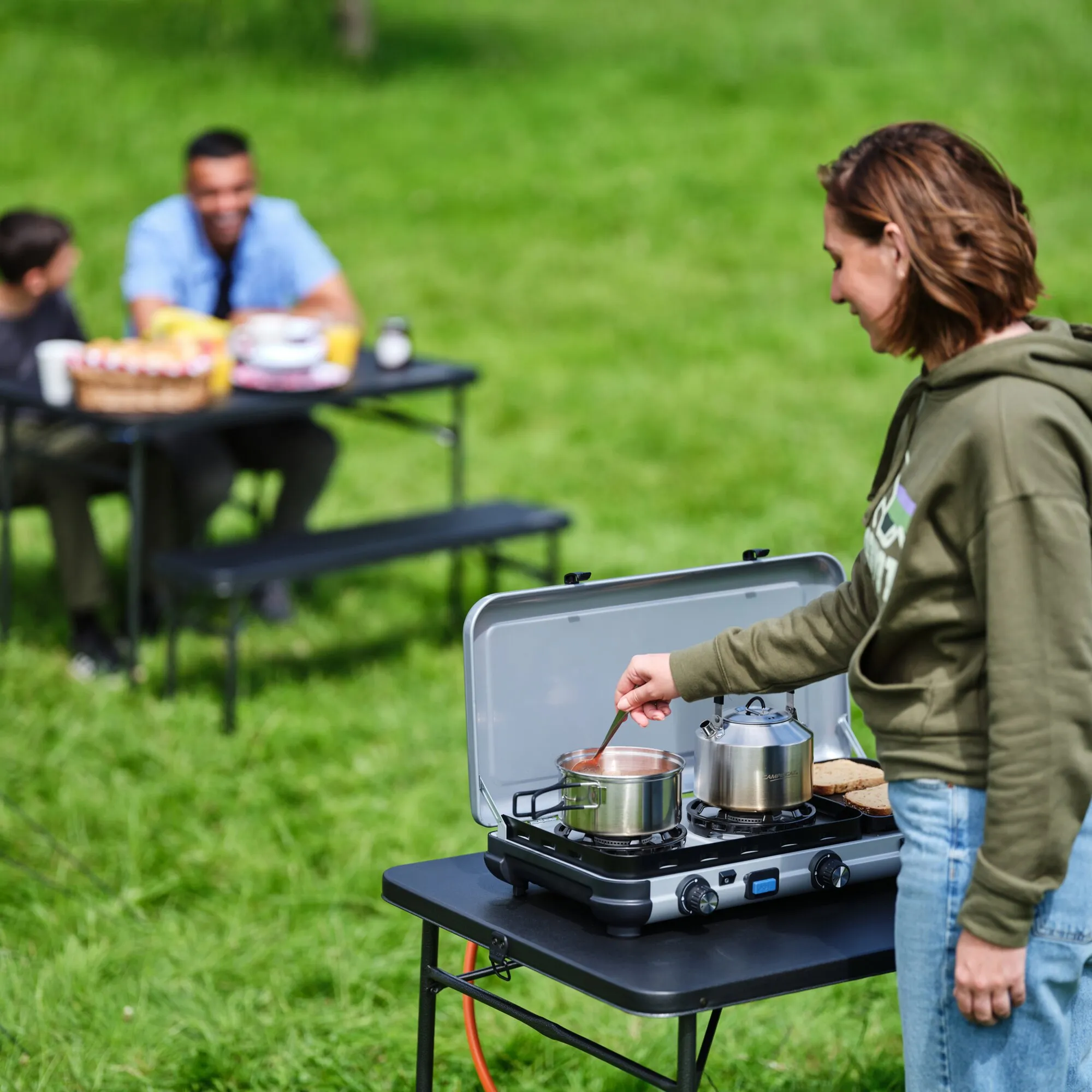 Zweiflammiger Gaskocher Camping Kitchen 2 MULTI-COOK beim Kochen im Freien: Eine Frau rührt im Topf, daneben steht ein Wasserkessel