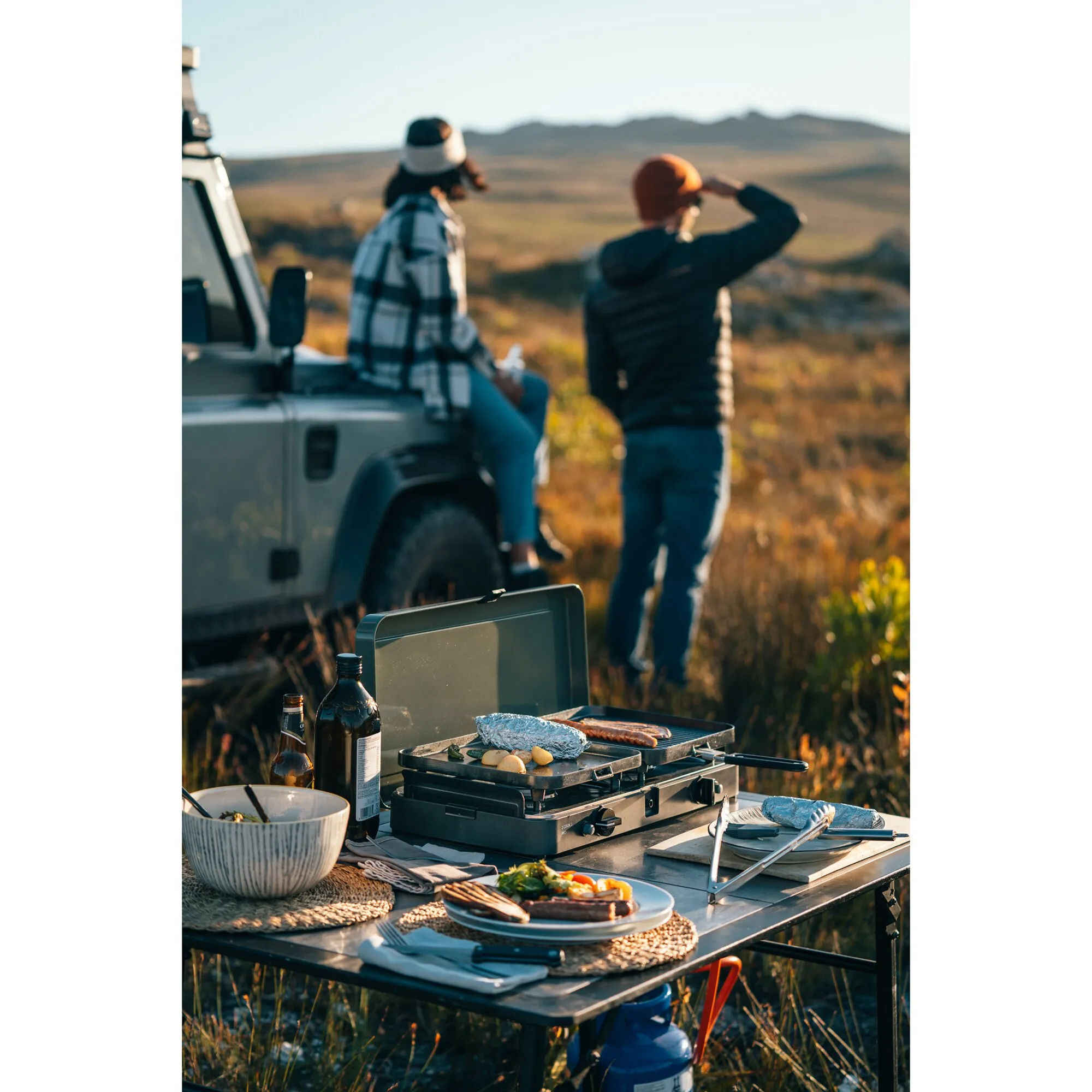 Schwarzer zweiflammiger Gaskocher 2 Cook 3 Pro Stove beim Outdoor-Kochen mit Grillplatten, Würstchen und Gemüse auf einem Campingtisch