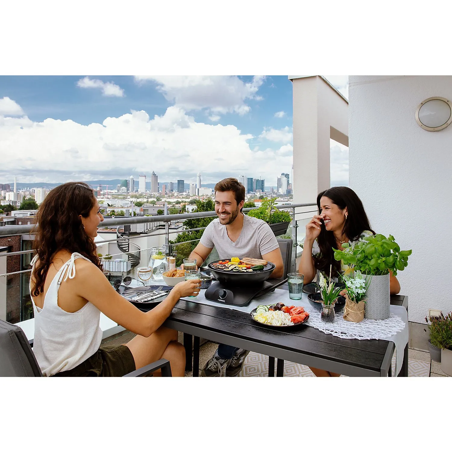 Drei Personen grillen auf einem Balkon am Tischgrill 360 Grill CV mit Gemüse und Fleisch vor einer Stadtkulisse