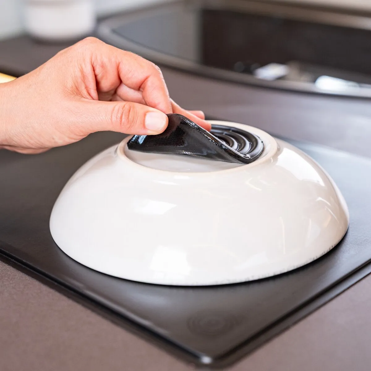 Hand hebt ein schwarzes MagUp-Pad, ein flexibles magnetisches Nachrüst-Pad, von einer weißen runden Unterlage auf einer Kochplatte ab