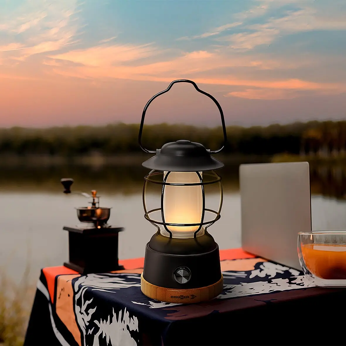 Lyss Akku-Campinglampe mit Glasbehälter und Metallkäfig, beleuchtet, steht auf Tischdecke am See bei Sonnenuntergang, links Kaffeemühle, rechts Laptop