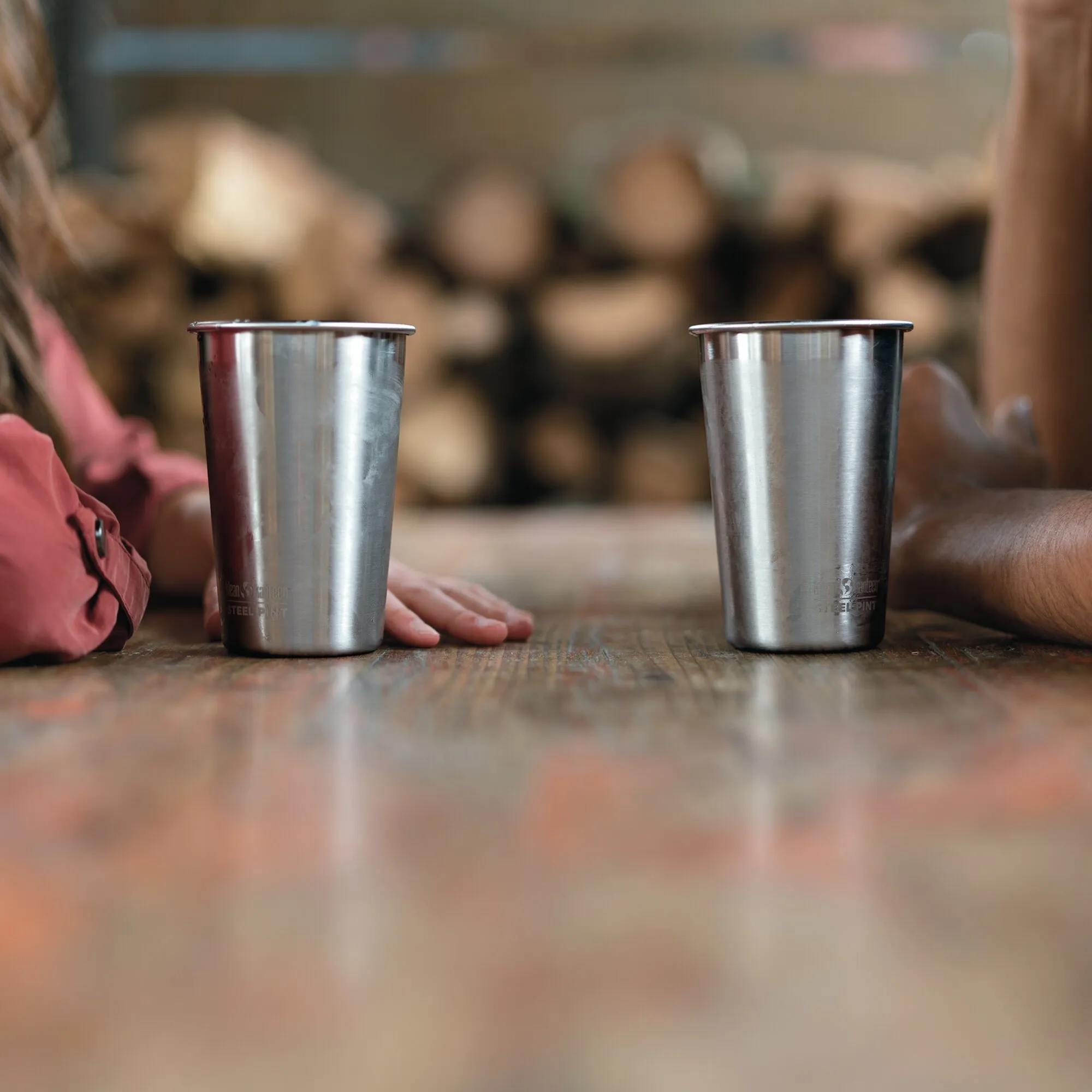 Zwei silberne Trinkbecher aus Edelstahl stehen auf einem Holztisch in einer geselligen Umgebung; Aufdruck Steel Pint sichtbar
