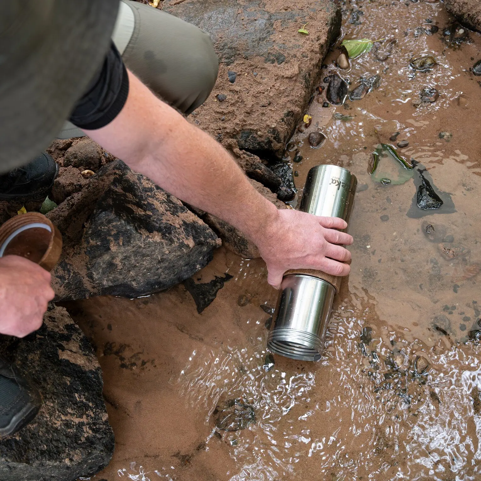 Eine Person hält einen glänzenden Edelstahl-Wasserfilter Travelbuddy über schlammigem Wasser mit Steinen, das Gewinde am Ende ist sichtbar