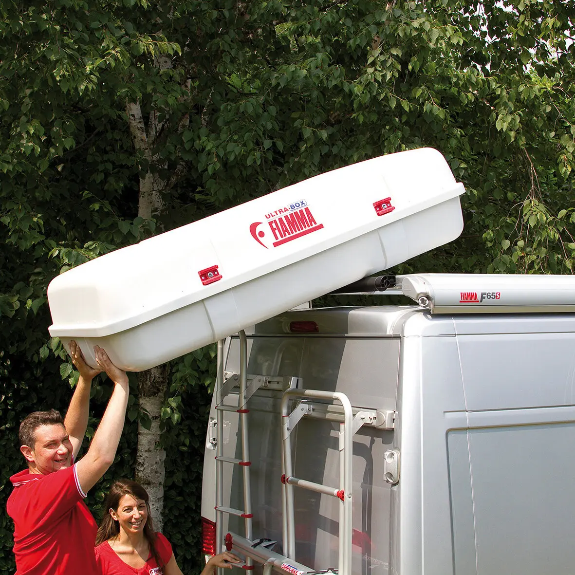 Zwei Personen heben eine weiße, längliche Dachbox mit roten Verriegelungen auf das Dach eines Transporters; am Fahrzeug ist eine FIAMMA Roller Roof Rail sichtbar