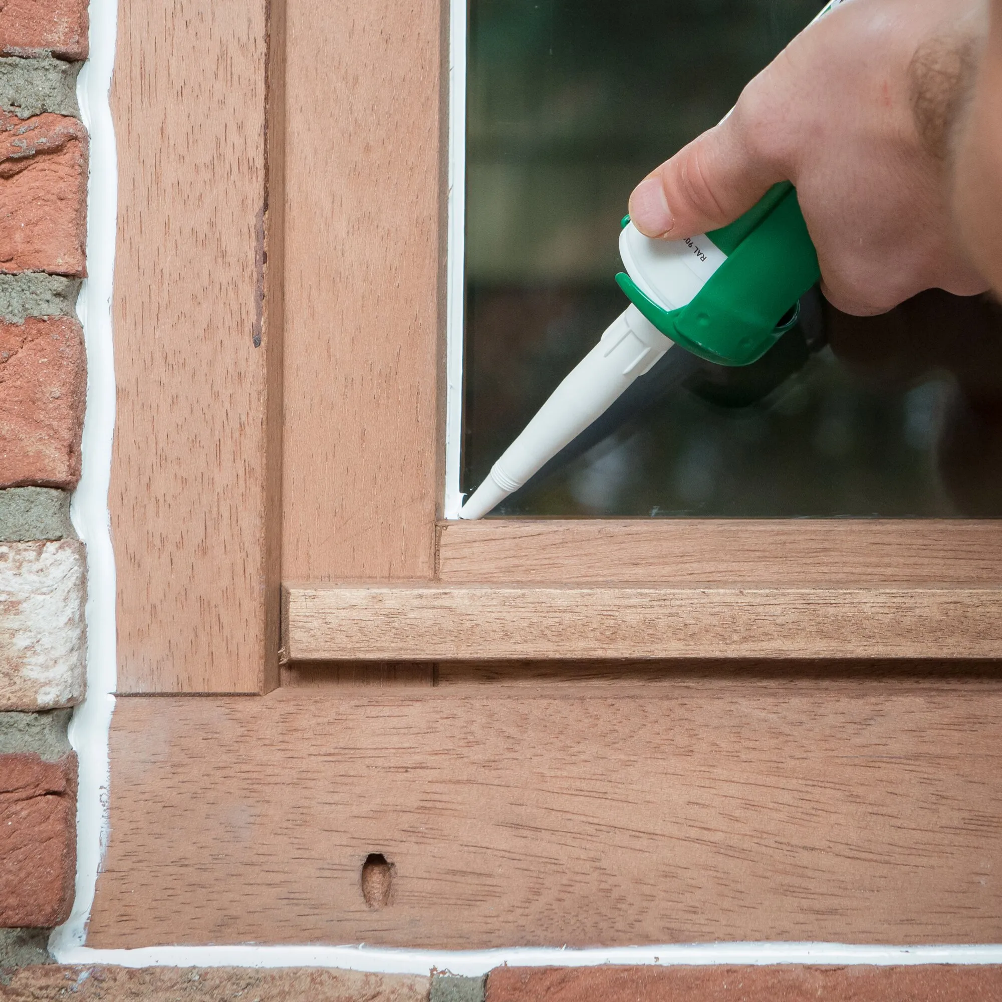Hand trägt Dichtstoff XSEAL EXTREME mit Kartuschenpistole entlang des Holzfensterrahmens neben einer Ziegelwand auf
