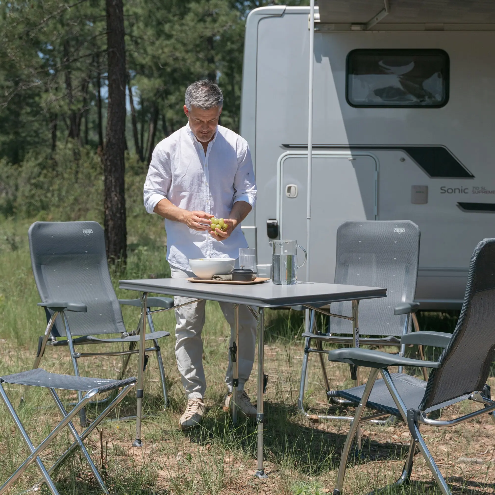 Person in hellem Hemd steht am Campingtisch Ligero 101 cm im Freien neben einem Wohnmobil, auf dem Tisch Gläser, eine Karaffe und Obst; graue Klappstühle ringsum, Wald im Hintergrund