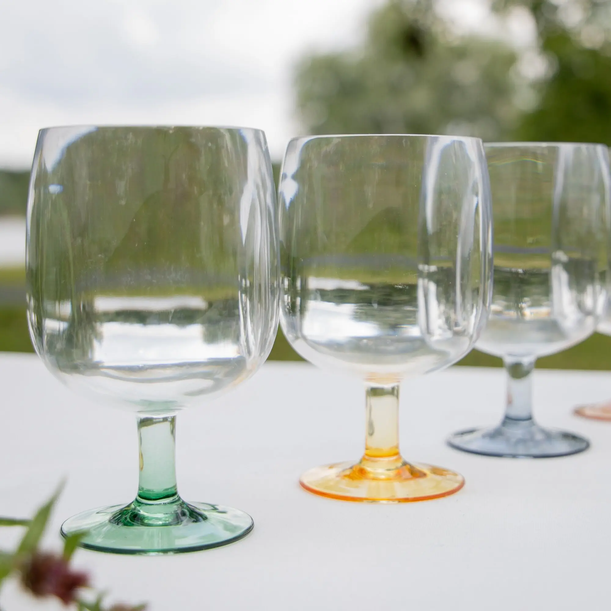 Nahaufnahme mehrerer bunter Weingläser mit farbigen Stielen (grün, blau, orange) auf weißen Tisch vor unscharfem Gartenhintergrund