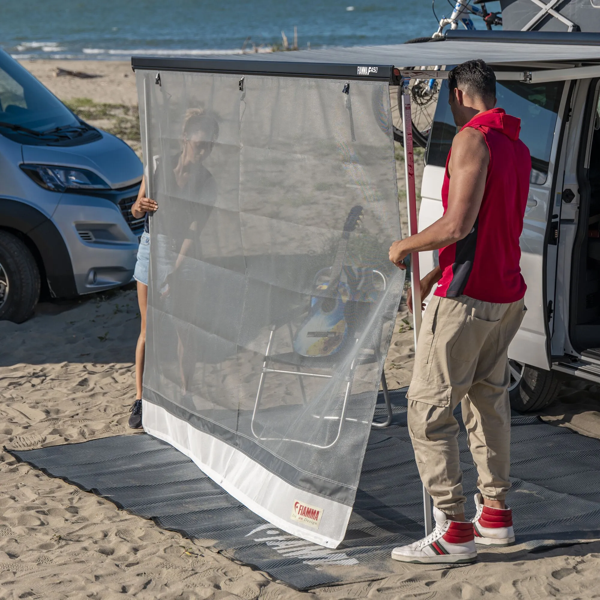 Zwei Personen montieren die graue Sun View XL 400 Vorderwand an einem Wohnmobil am Strand; das Fiamma-Logo ist sichtbar