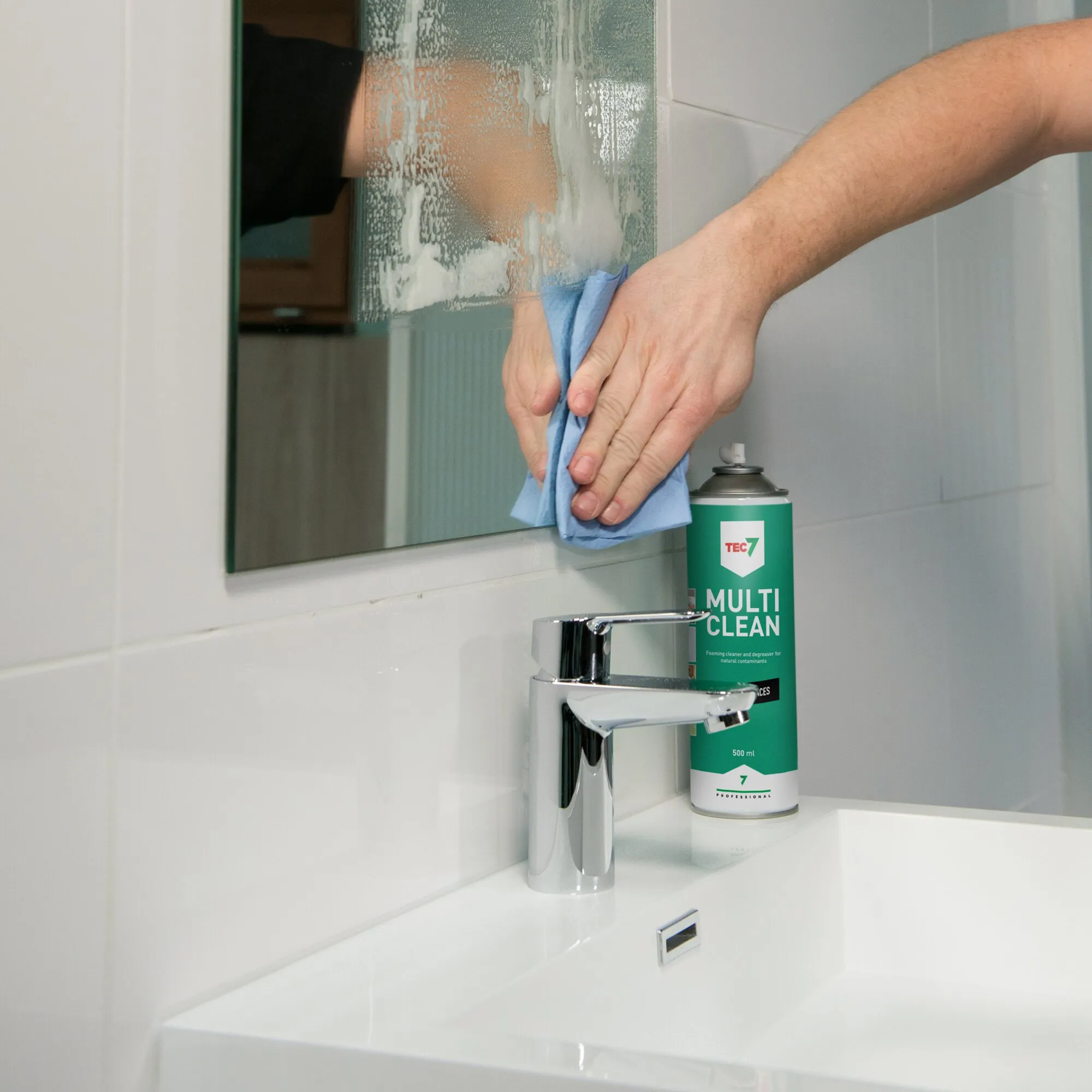 Hand wischt mit blauem Tuch Schaum von einem Badezimmerspiegel; daneben steht die grüne Dose TEC7 MULTI CLEAN Schaumreiniger