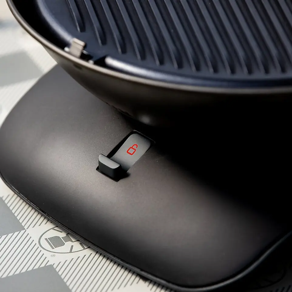 Nahaufnahme eines runden Tischgrills mit gerippter Grillfläche und einem seitlichen Gehäuseschalter mit rotem Schloss-Symbol