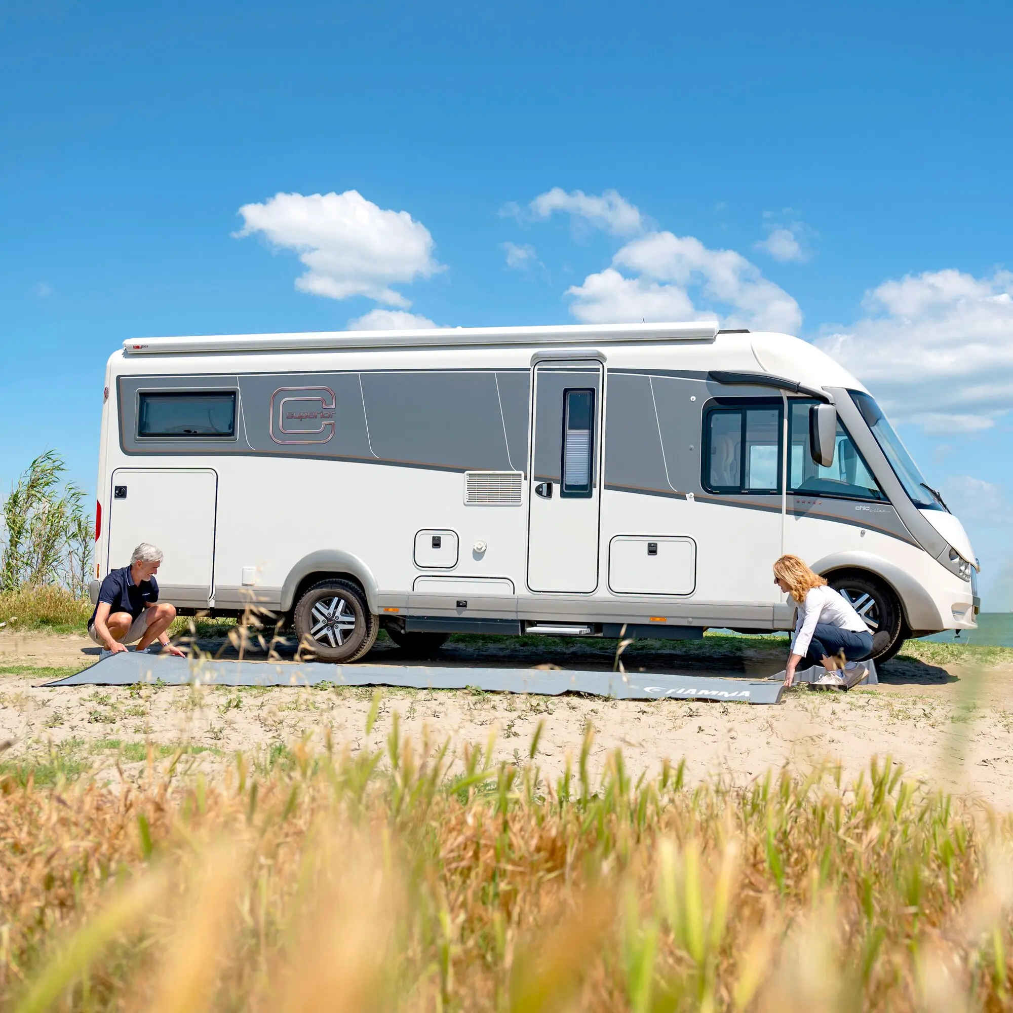 Patio-Mat Zeltteppich, 290 cm, wird vor einem Wohnmobil am Strand ausgerollt; zwei Personen breiten ihn flach aus, Gras im Vordergrund, blauer Himmel