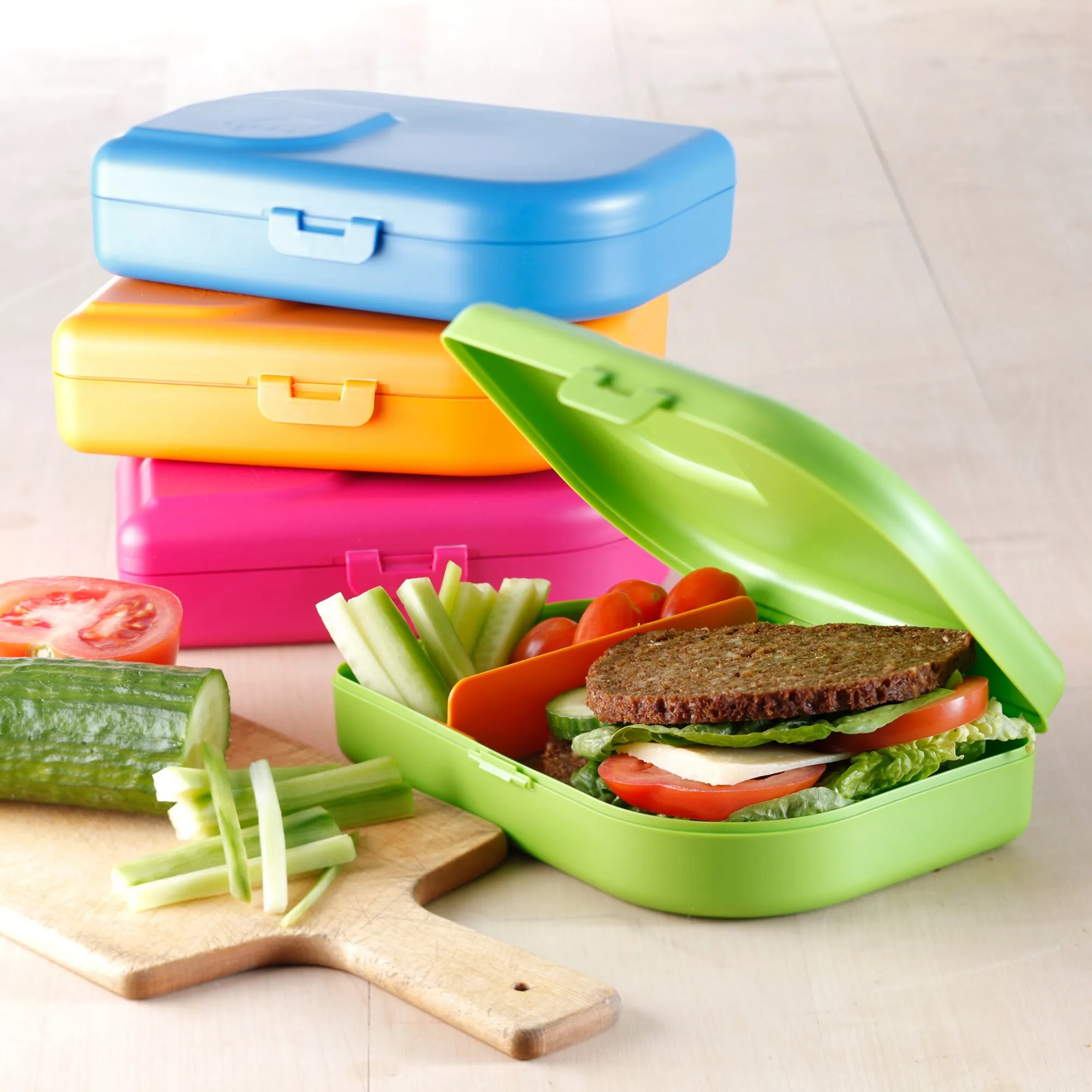 Blaue Brotbox mit Verschluss, gestapelt auf orangefarbenen und pinken Boxen neben einer geöffneten grünen Brotbox mit Sandwich und Gemüse