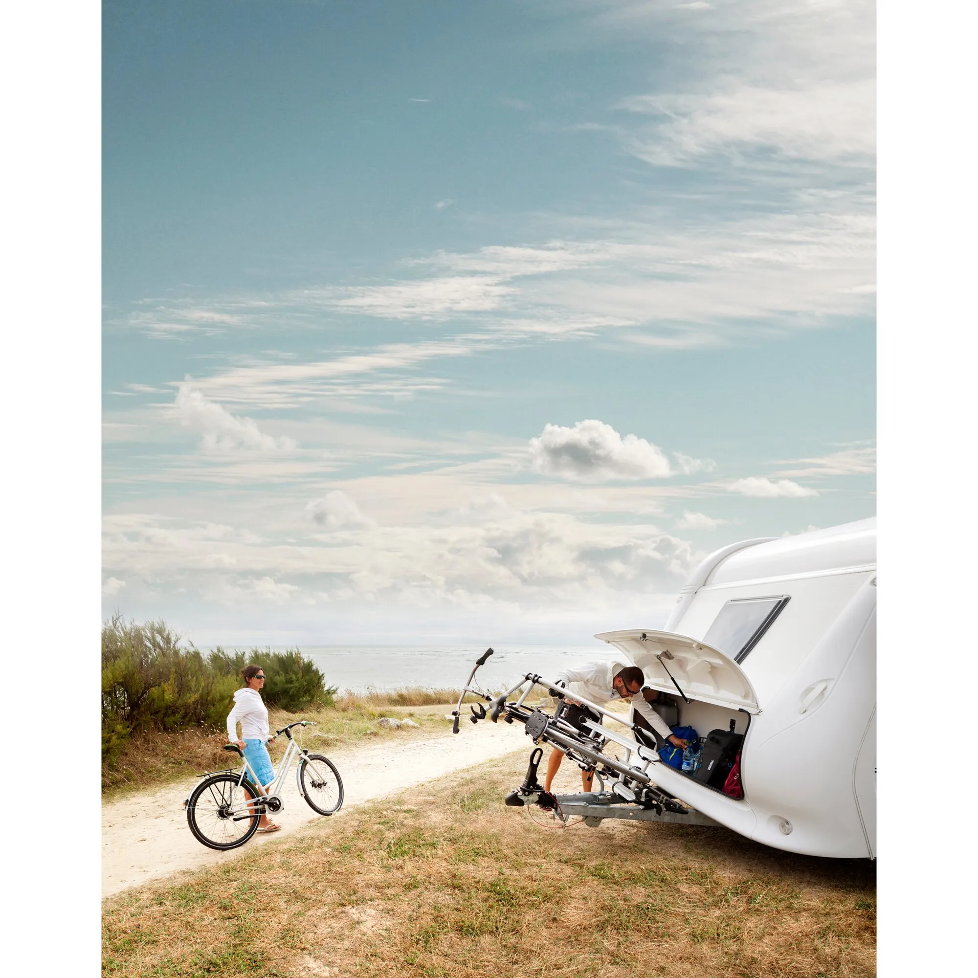 Weißer Wohnwagen am Meer mit Thule Caravan Superb XT Fahrradträger; ein Mann lädt Fahrräder, eine Frau steht daneben