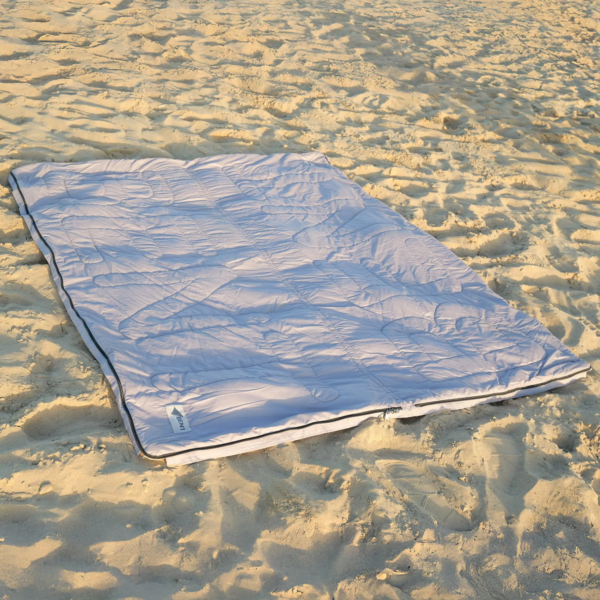 Hellgrauer, quiltierter Vanlife Schlafsack Bentcho auf Sandstrand, Reißverschluss mit dunkler Kante, Etikett am Rand sichtbar
