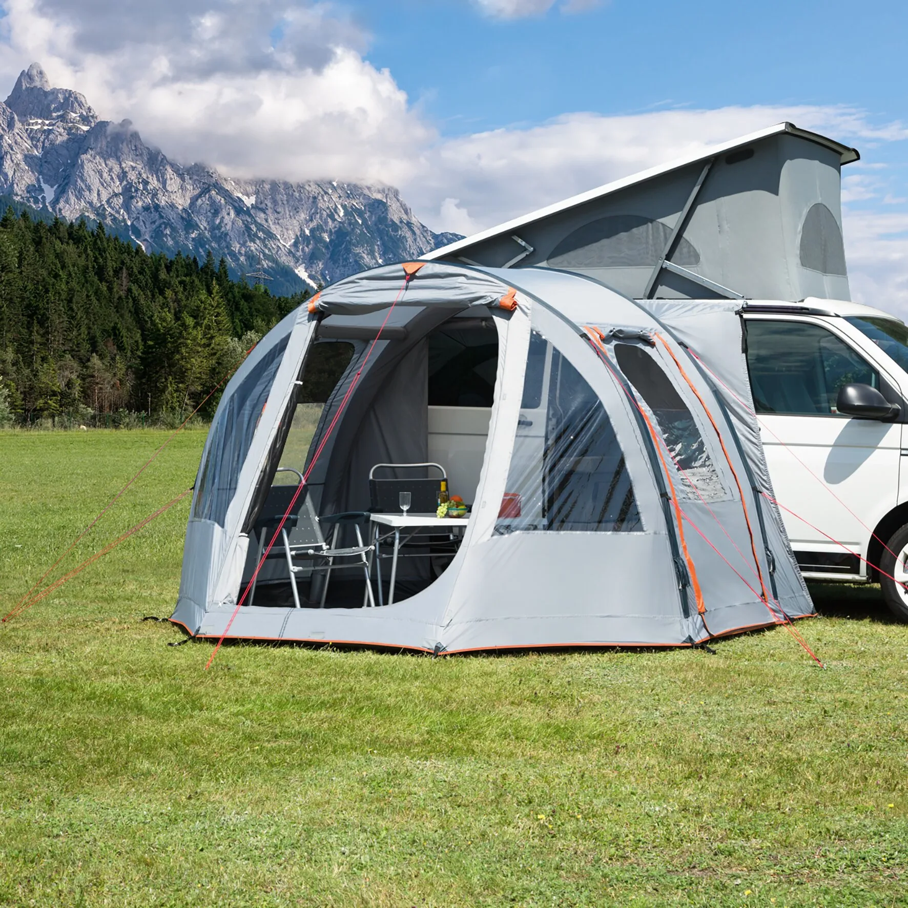 Busvorzelt Picco Air in Grau mit orangefarbenen Details, am weißen Campingbus aufgebaut, geöffnet mit Tisch und Stühlen vor Bergkulisse