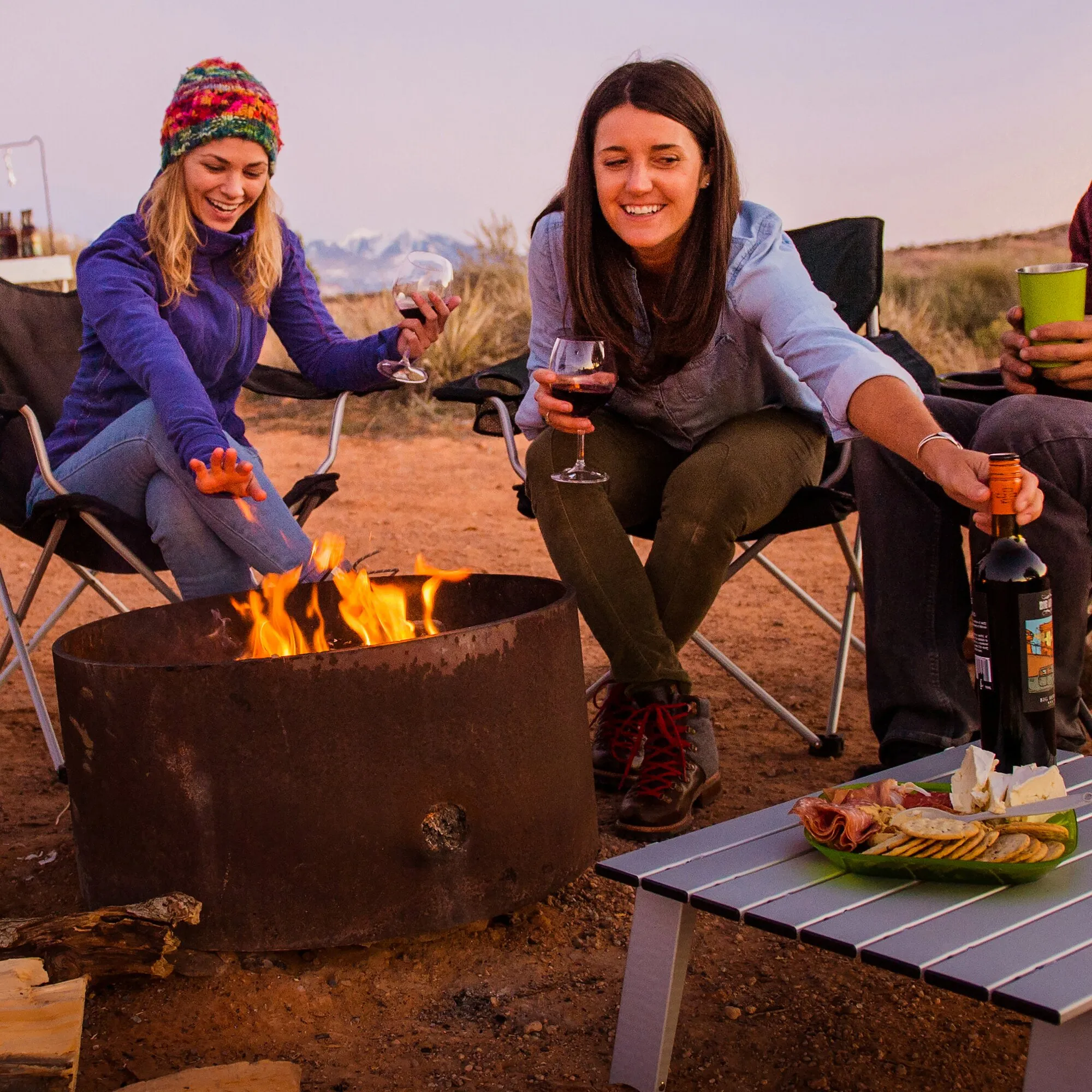 Beim Sitzen am Lagerfeuer halten zwei Frauen Rotwein in transparenten Nesting-Rotweingläsern