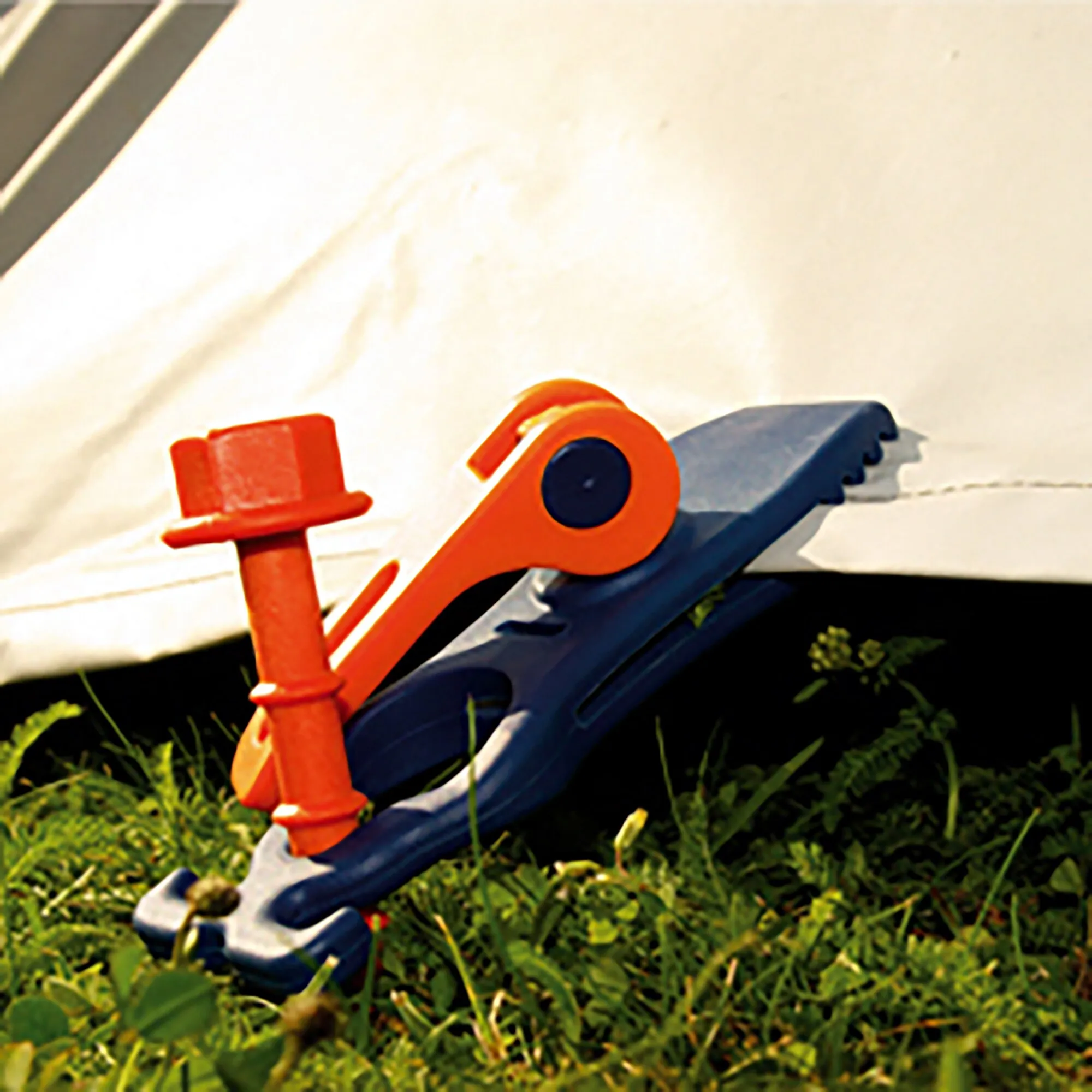 Orange-blaue Schnellspannklemme PP Crocodile 4 hält eine helle Plane an einer dunklen Kante im Gras fest