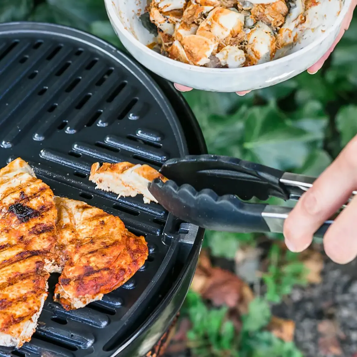 Grillzange aus Edelstahl, 280 mm, greift ein Stück gegrilltes Fleisch vom runden Grillrost; daneben eine Schale mit weiteren Fleischstücken, Hintergrund mit Grünpflanzen