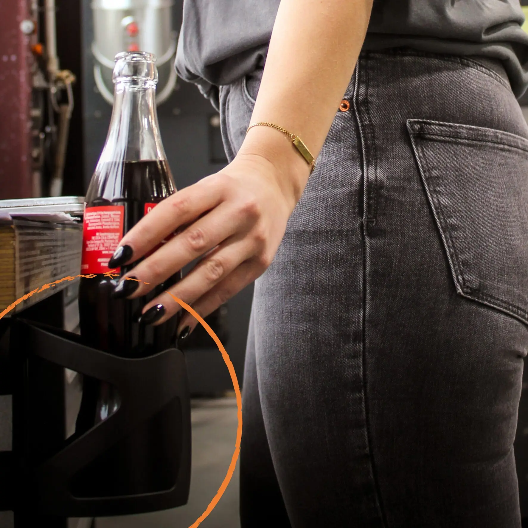 Person in grauer Jeans mit goldenem Armreif steht neben einer Magnethalterung UNI, in der eine Coca‑Cola Flasche steckt