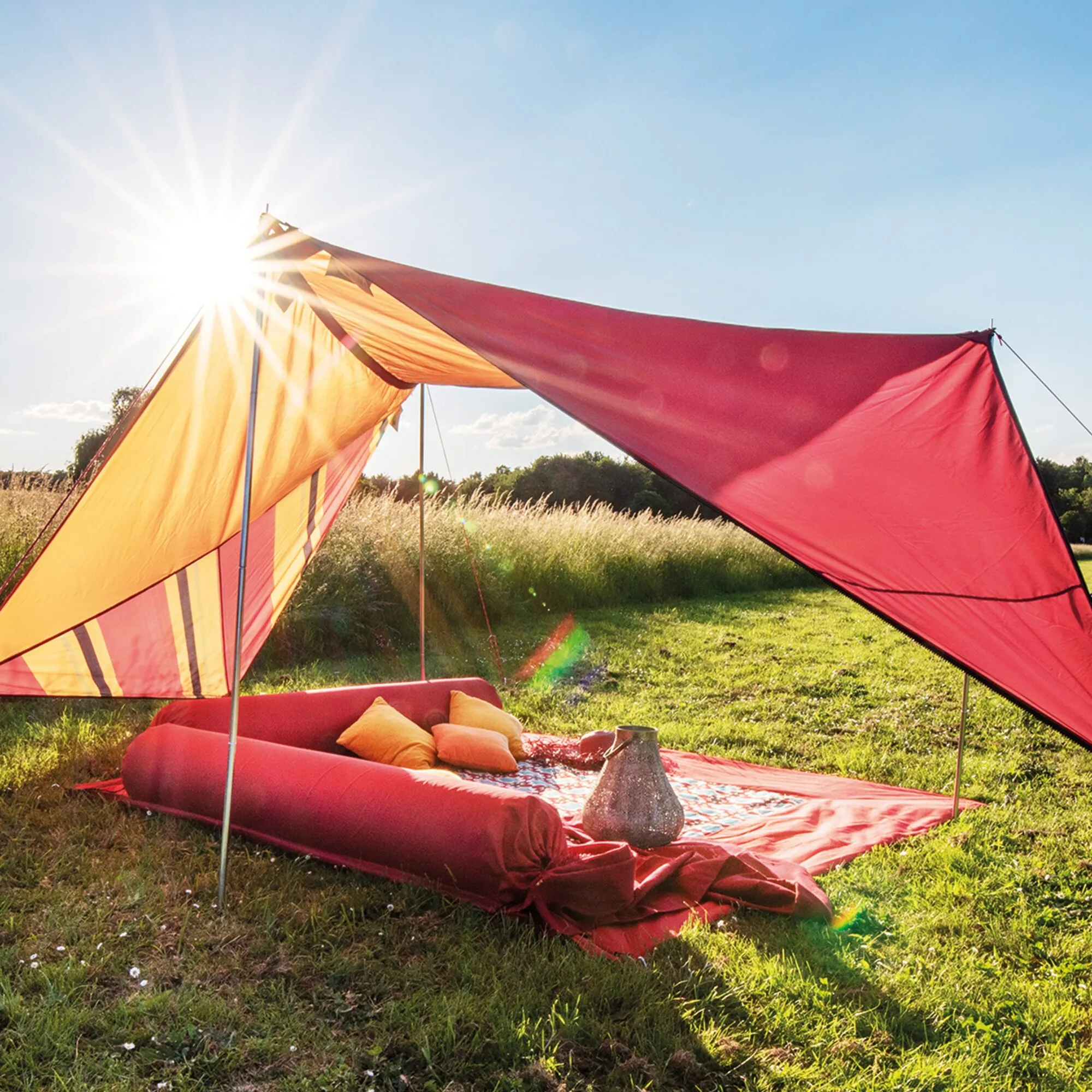Rotes und orangefarbenes Sonnensegel Zip Canvas Oriental auf einer Wiese, mit Sitzkissen und Picknickdecke unter blauem Himmel