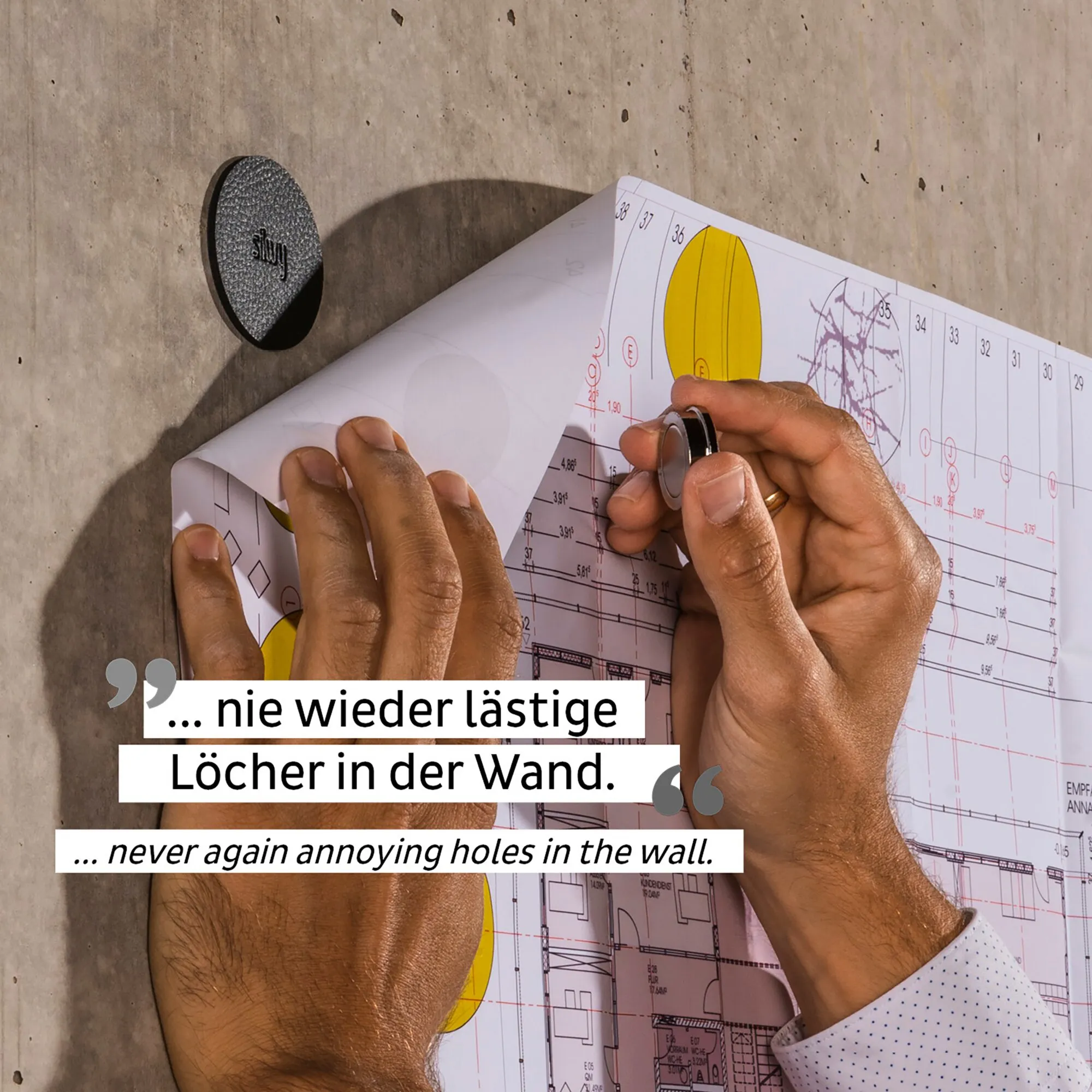 Schwarzer runder Magnet-Pin SMART an einer Betonwand, während ein weiterer Pin einen Bauplan ohne Bohrlöcher fixiert