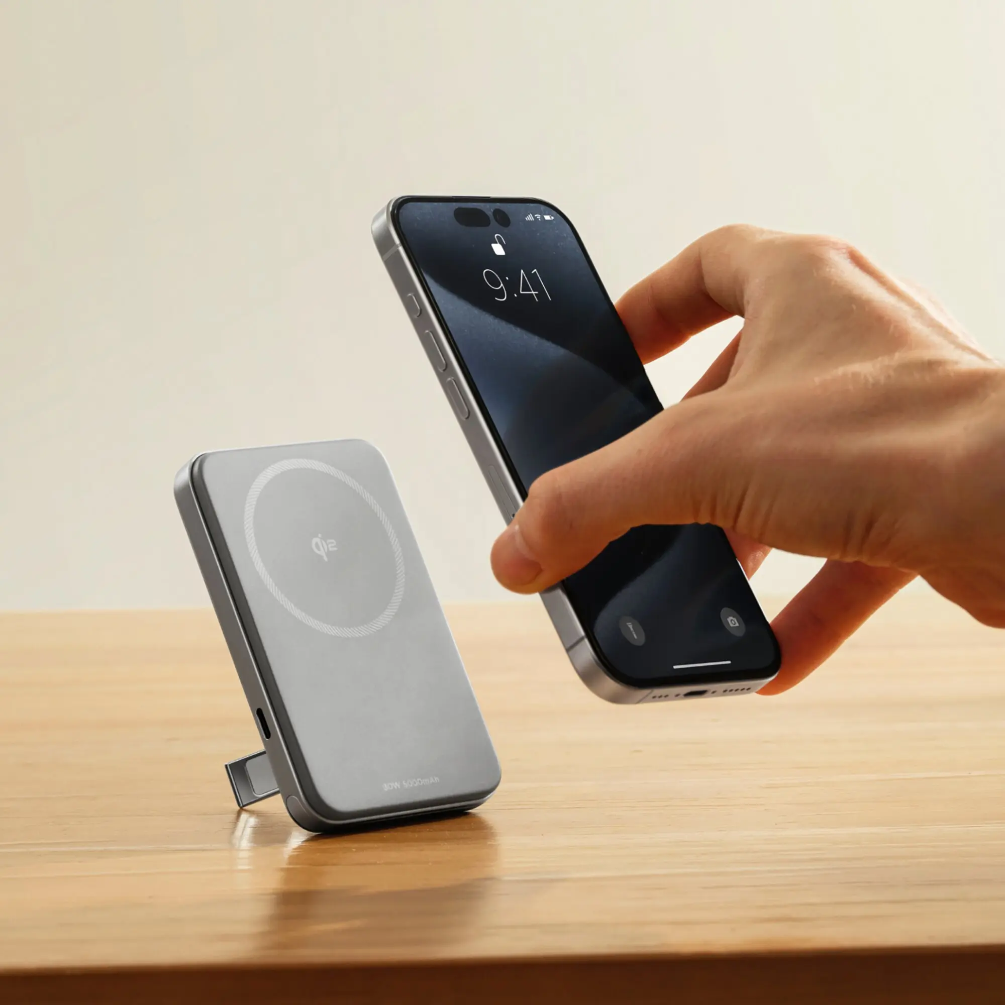 Hand hält ein Smartphone nahe an einer magnetischen Powerbank Rapid, 5000 mAh, daneben liegt eine graue Qi-Ladeplatte auf einem Holzuntergrund