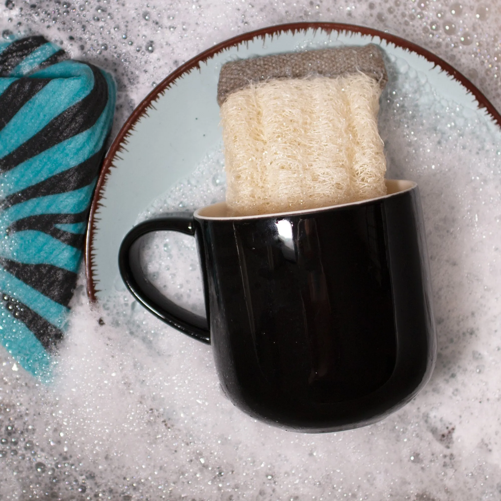Cremefarbener Luffa-Schwamm in einer schwarzen Tasse auf einer türkis gemusterten Keramikablage, umgeben von Seifenschaum