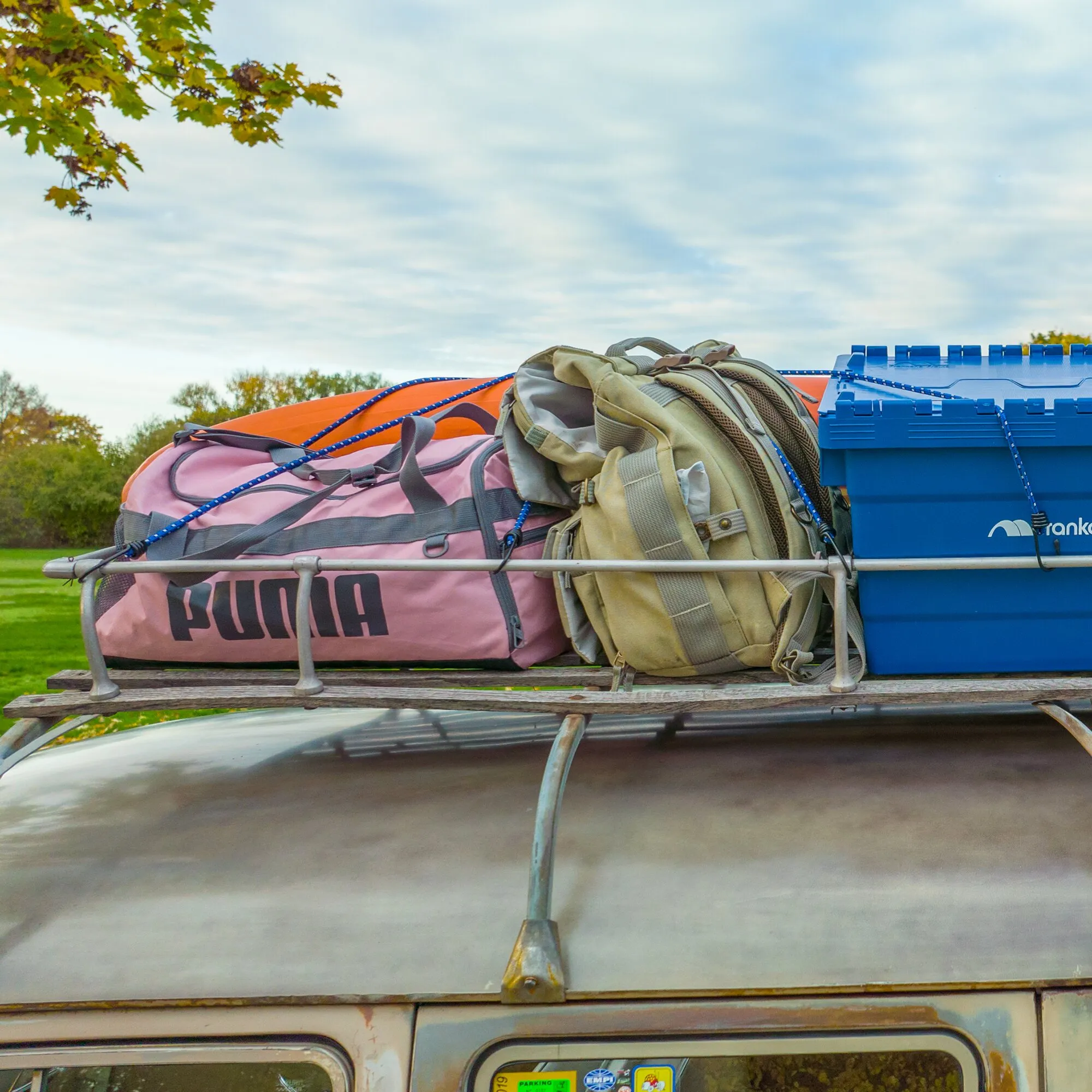 Blaue Gepäckspinne sichert eine pinke Tasche, einen Rucksack und eine blaue Transportbox auf dem Dachträger eines Fahrzeugs