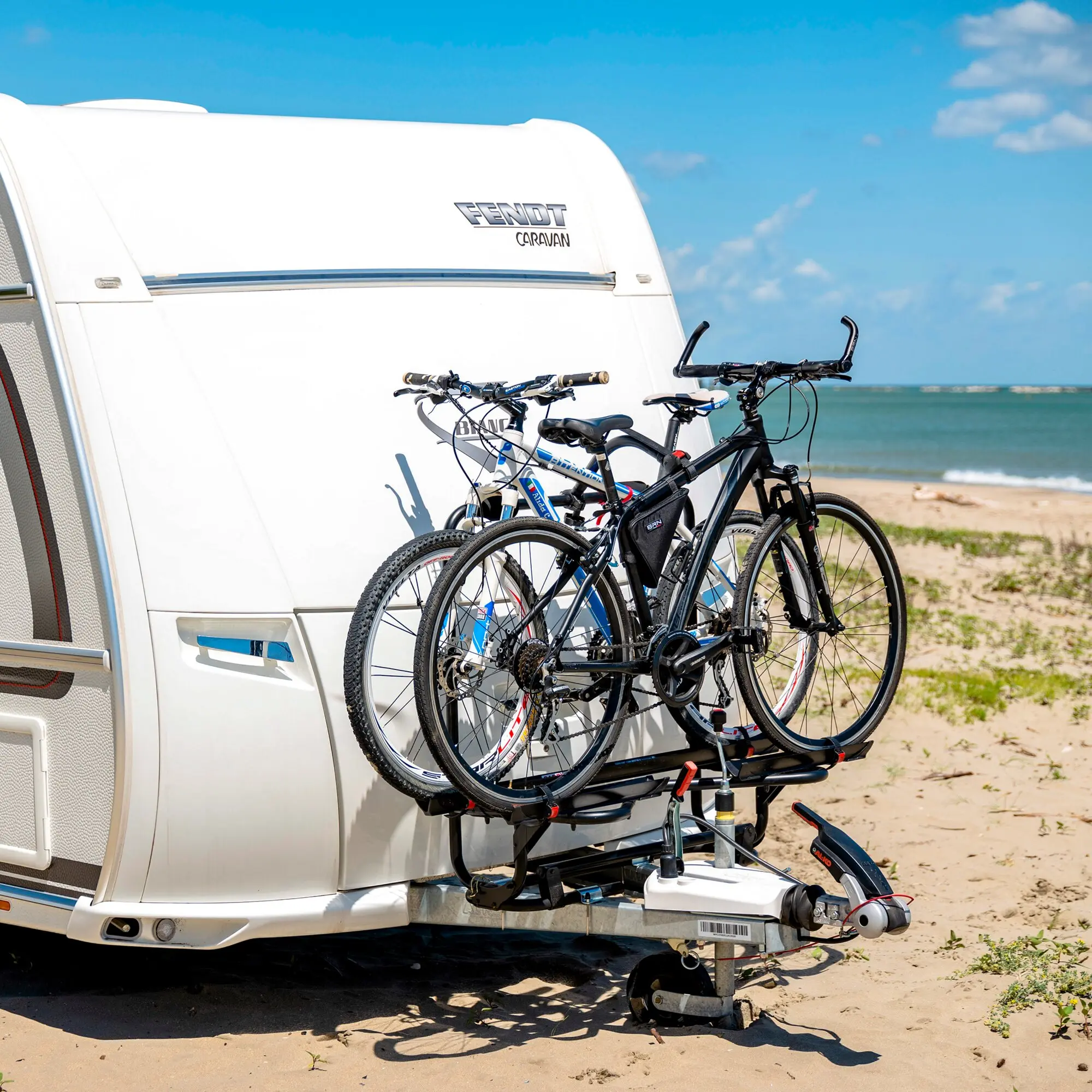 Rückseite eines weißen Caravans mit schwarzem Heckträger Carry-Bike Caravan XL A Pro 300, zwei Fahrräder montiert, Strand im Hintergrund
