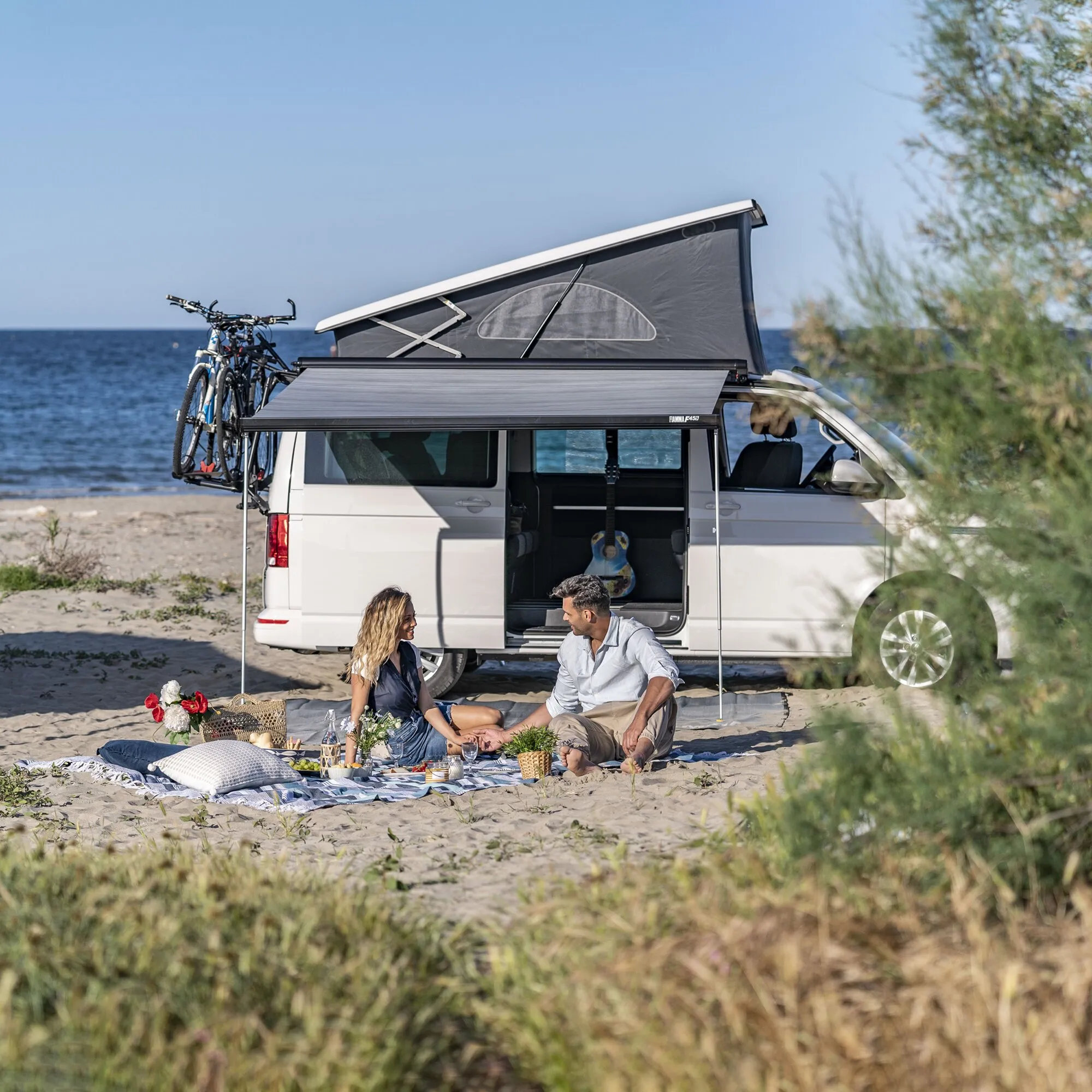 Schwarze Fiamma F45s-Markise, 308 cm, am weißen VW T5/T6 Multivan ausgefahren; ein Paar sitzt am Strand darunter