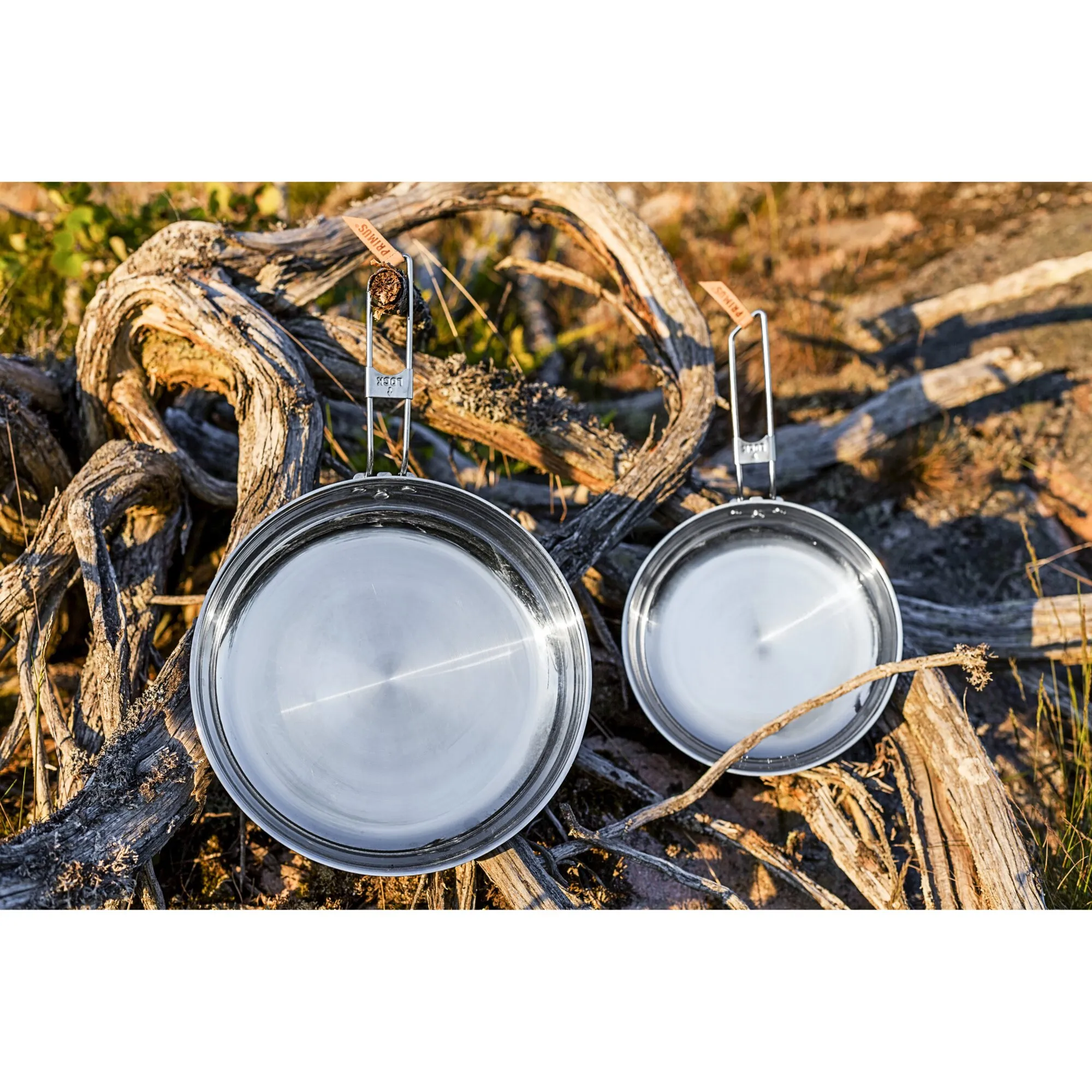 Zwei silberne Campfire-Pfannen aus Edelstahl mit klappbaren Drahtgriffen liegen auf trockenem Holz in der Natur