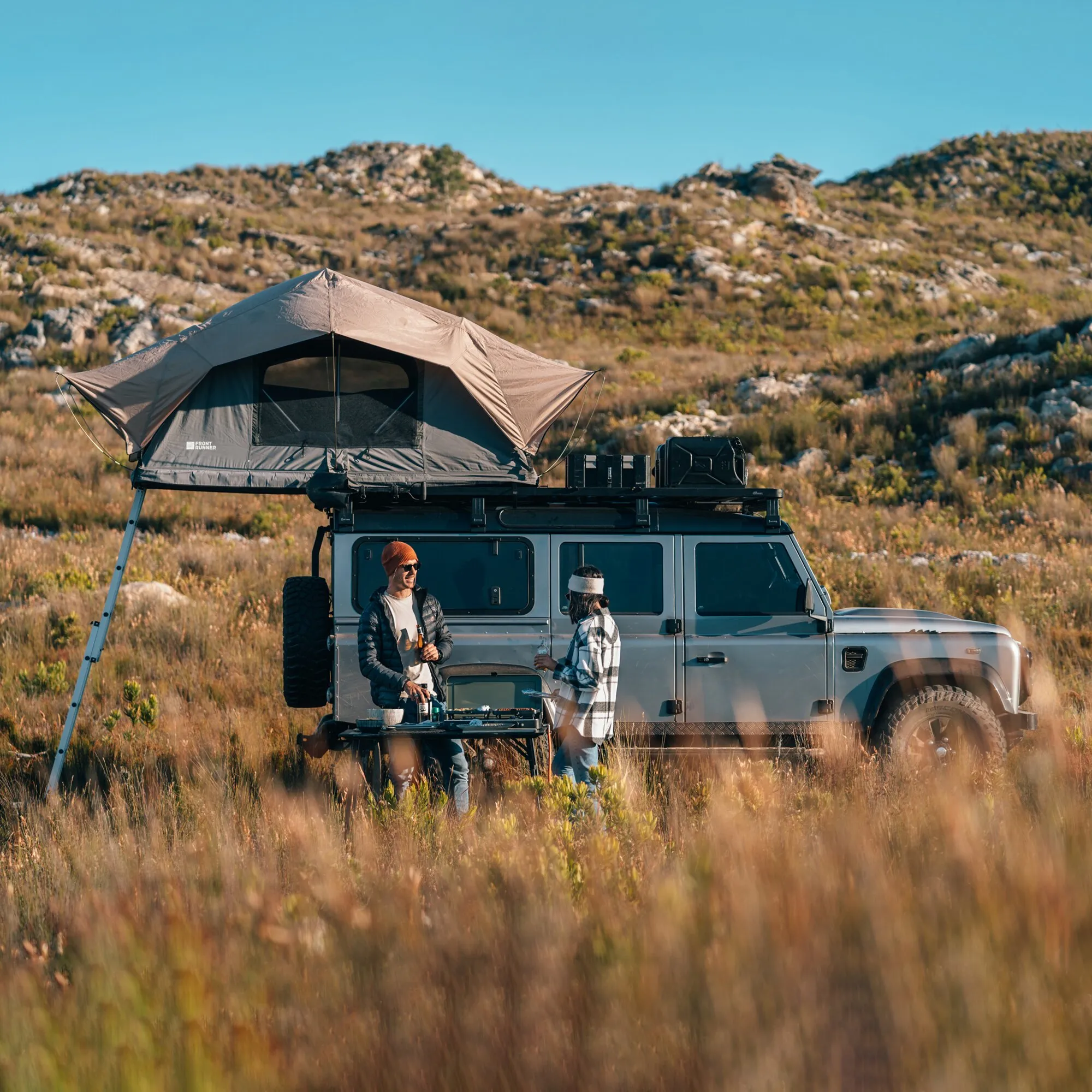 Zweiflammiger Gaskocher 2 Cook 3 Pro Stove beim Kochen mit Töpfen auf einem Campingtisch neben einem Geländewagen
