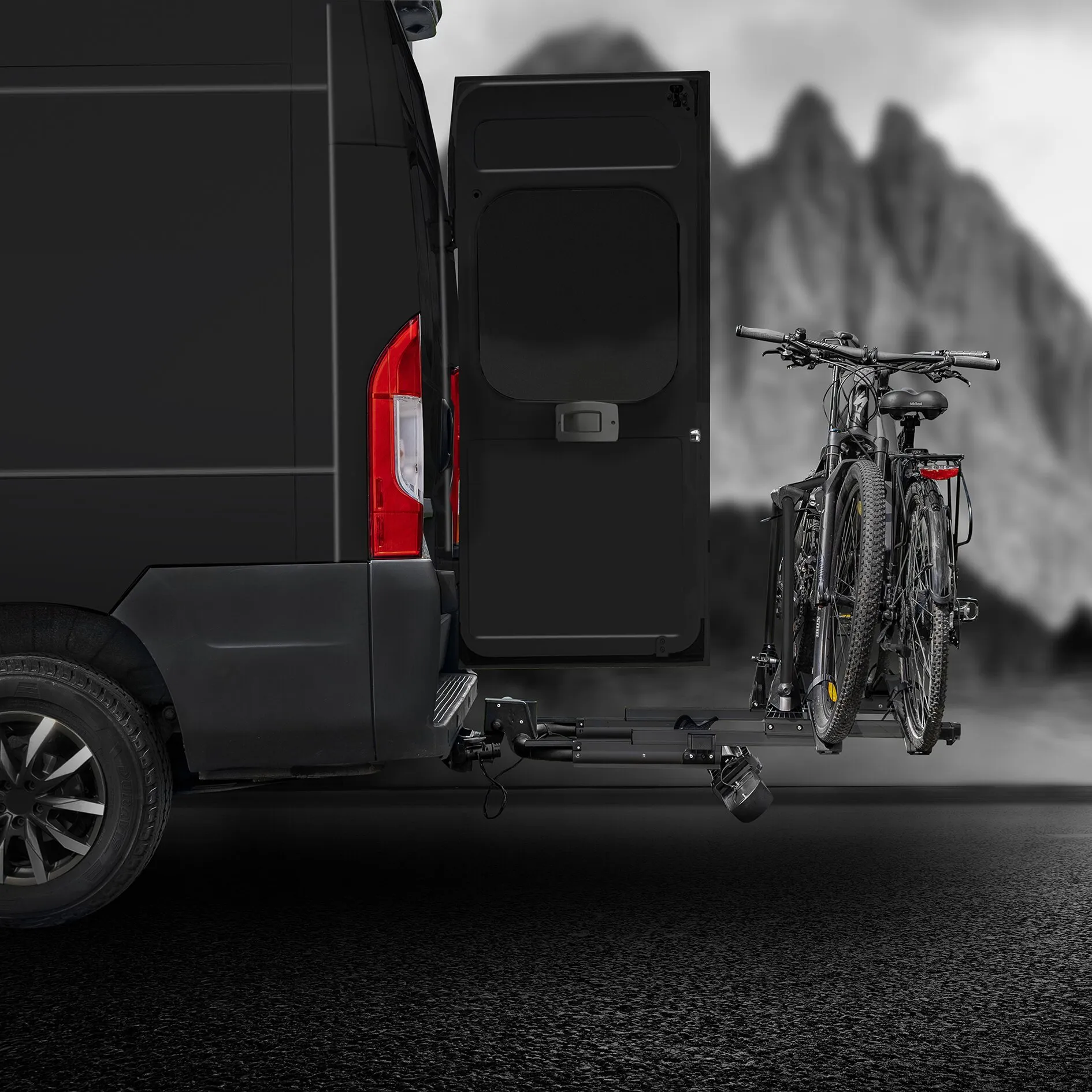 Schwarzer SD260 Fahrradträger an der Anhängerkupplung eines schwarzen Vans, zwei Fahrräder beladen, Heck geöffnet, Berglandschaft im Hintergrund