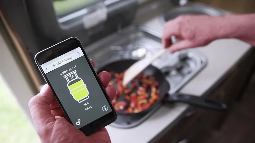 Smartphone mit der App E-Gaslevel zeigt 85 Prozent beziehungsweise 8,5 Kilogramm Gasfüllstand, während auf einem Gaskocher gekocht wird