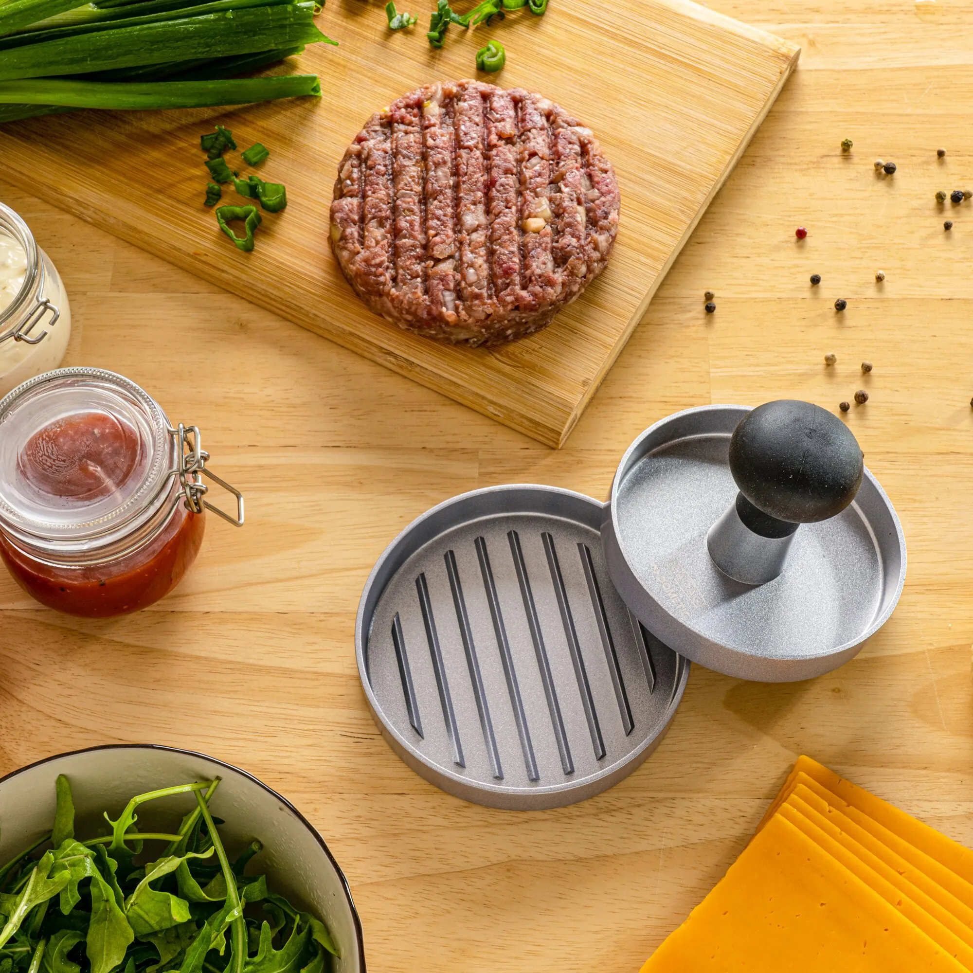 Graue Hamburgerpresse mit geriffelter Grillplatte und schwarzem Griff neben rohem Burgerpatty, Käse, Salat und Saucenglas