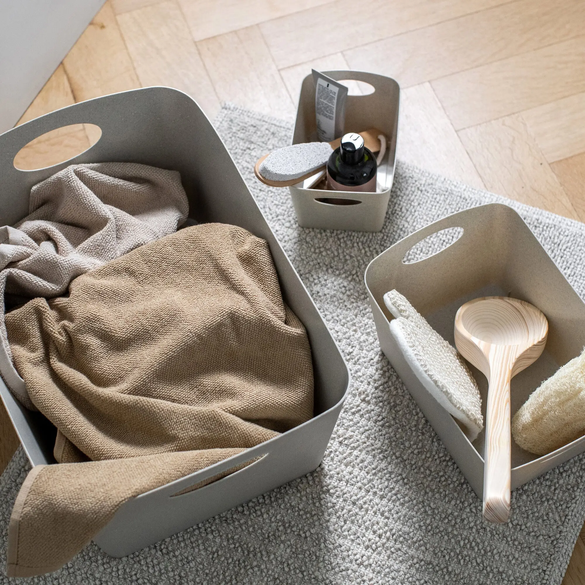 Sandfarbene Aufbewahrungsbox BOXXX M mit Griffen auf hellgrauem Teppich; innendrin beige Stoffe und eine Pumpspenderflasche sichtbar