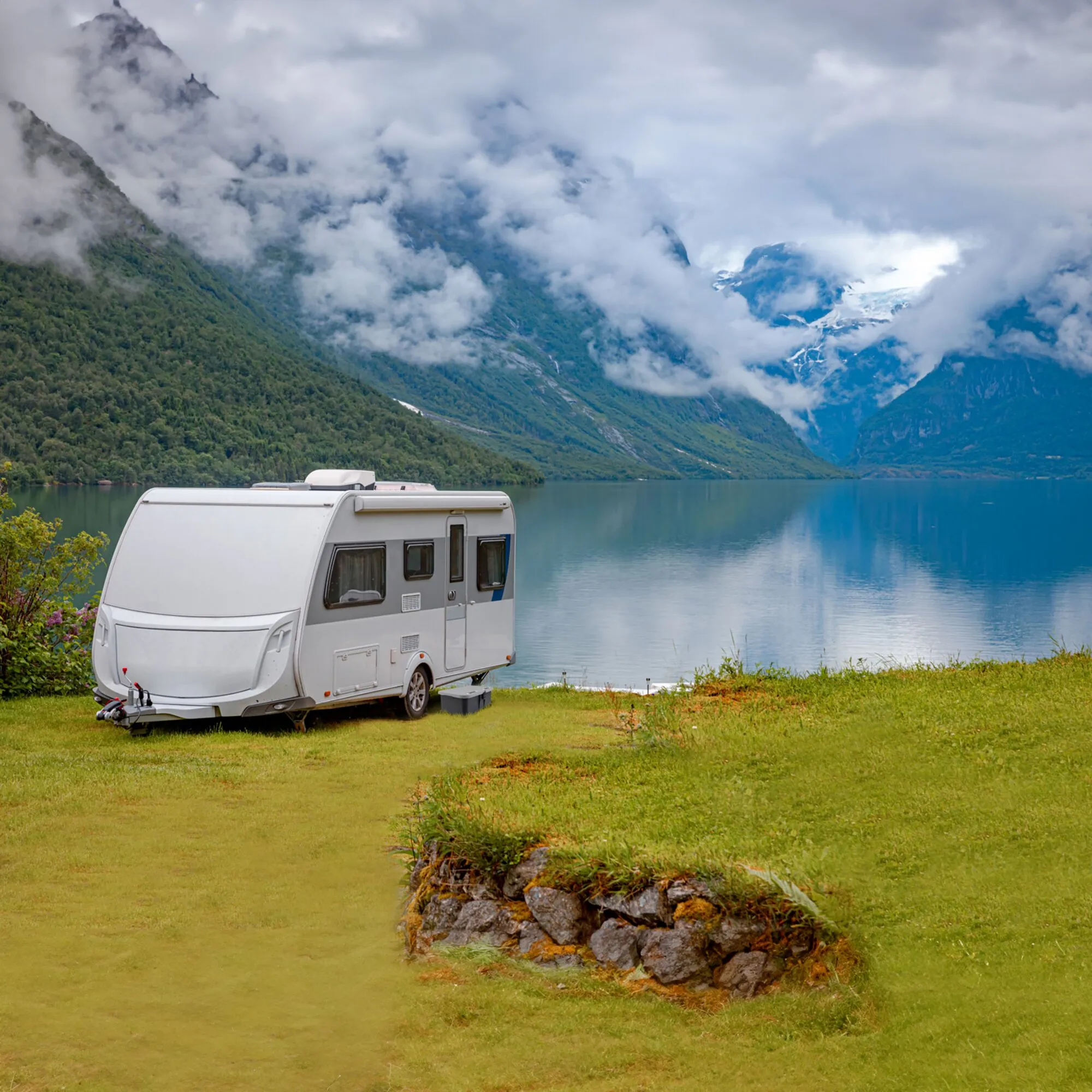 Weißer Wohnwagen am See mit grauer Trittstufe Boxstep NG vor dem Einstieg, umgeben von Bergen und Wolken