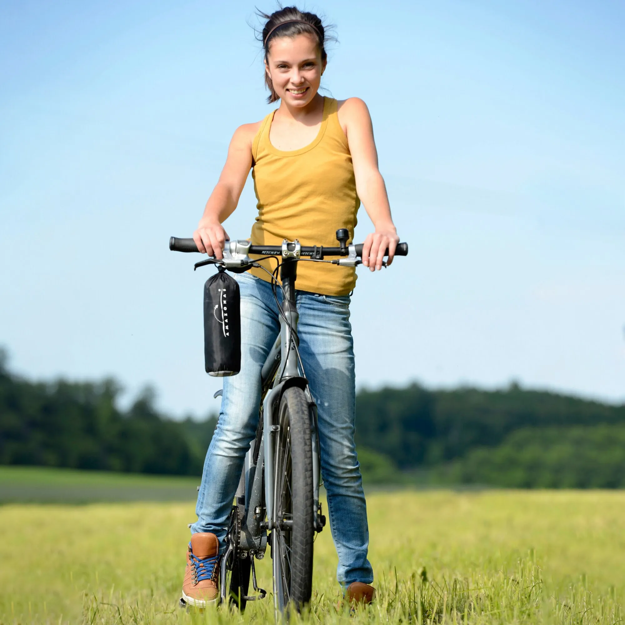 Frau mit Fahrrad auf einer Wiese; am Lenker hängt die zusammengerollte Picknickdecke Ultra-Light in schwarzer Tragtasche